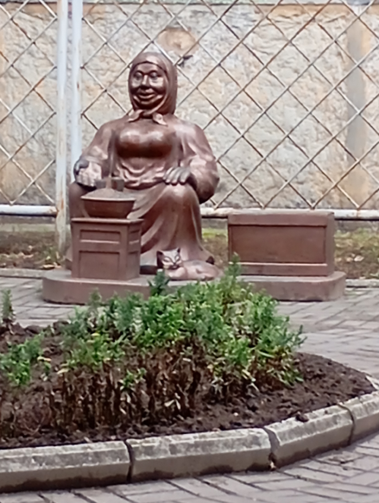 The photograph shows a bronze sculpture of a seated woman wearing a headscarf and a long dress. She is depicted with a smiling expression. In front of her is a small, stool-like object with a bowl on top. To her right, a small cat sculpture is visible. Behind the woman, there is a low, rectangular structure. The sculpture is situated outdoors, with a textured fence or wall in the background. In the foreground, there is a planter bed with green foliage and dark soil, bordered by concrete edging and paving stones. This scene is located in the city of Horishni Plavni, Ukraine.
