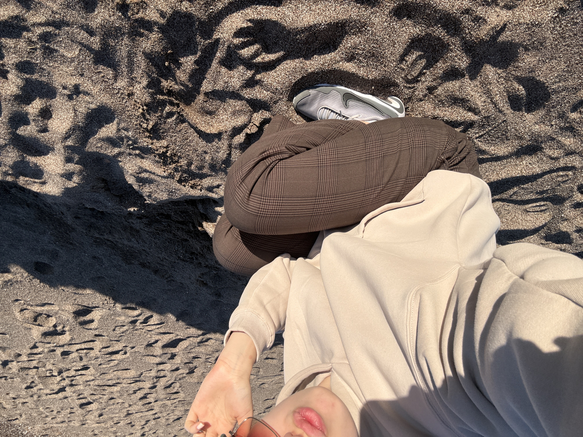 The image presents a top-down, partial view of a person lying on a dark sand beach in Boreti, Montenegro. The individual is dressed in a light beige hooded sweatshirt and brown plaid trousers. Their face is partially visible in the bottom left, showing pink lips and a hand holding dark-lensed sunglasses.
A single grey and black Nike sneaker, identifiable by its prominent swoosh logo and brand text, rests on the sand near the person's legs. The sand itself is a striking feature, appearing very dark, almost black, with individual grains sparkling brightly under direct sunlight, indicating a coarse texture and possibly unique mineral content. Strong, elongated shadows are cast across the sand, suggesting a sunny day, likely late morning or afternoon. The person's relaxed posture implies they are enjoying the warm, sunny weather, possibly taking a self-portrait from their reclining position. The overall scene conveys a calm and casual atmosphere on a distinctive beach.