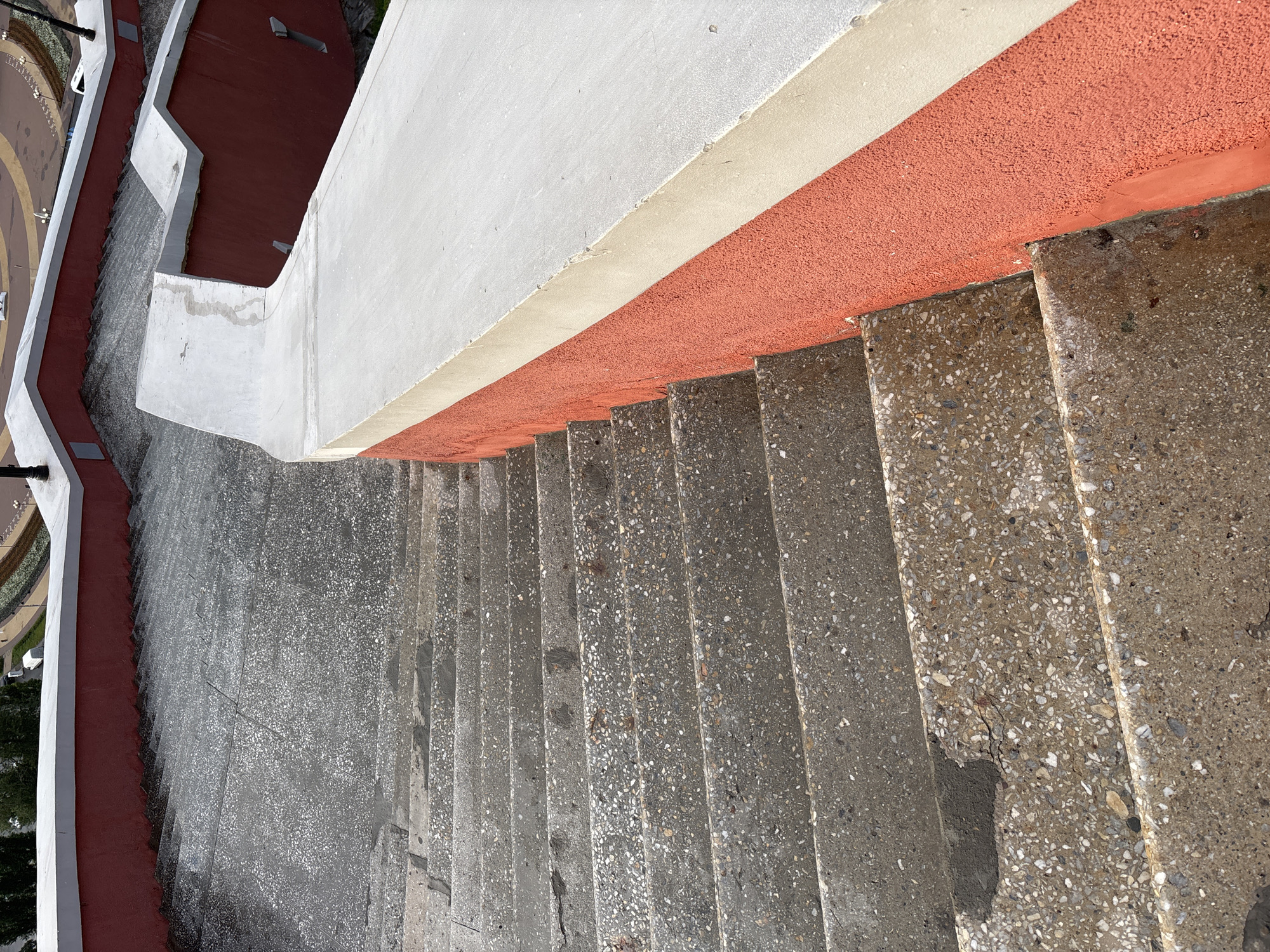 The image shows a wide, outdoor staircase constructed of textured concrete with visible aggregate. The staircase descends from the viewer's perspective, with multiple steps clearly defined. Along the right side of the stairs, there is a bright red wall with a thick, pale beige concrete railing on top. The railing curves along the stairs. In the upper portion of the image, a grey concrete surface with visible cracks is visible, indicating the top of the staircase or a landing. The overall impression is of a public outdoor architectural feature. The specific location in Nizhny Novgorod, Russia, is not discernible from the image itself. There are no people or specific actions depicted.