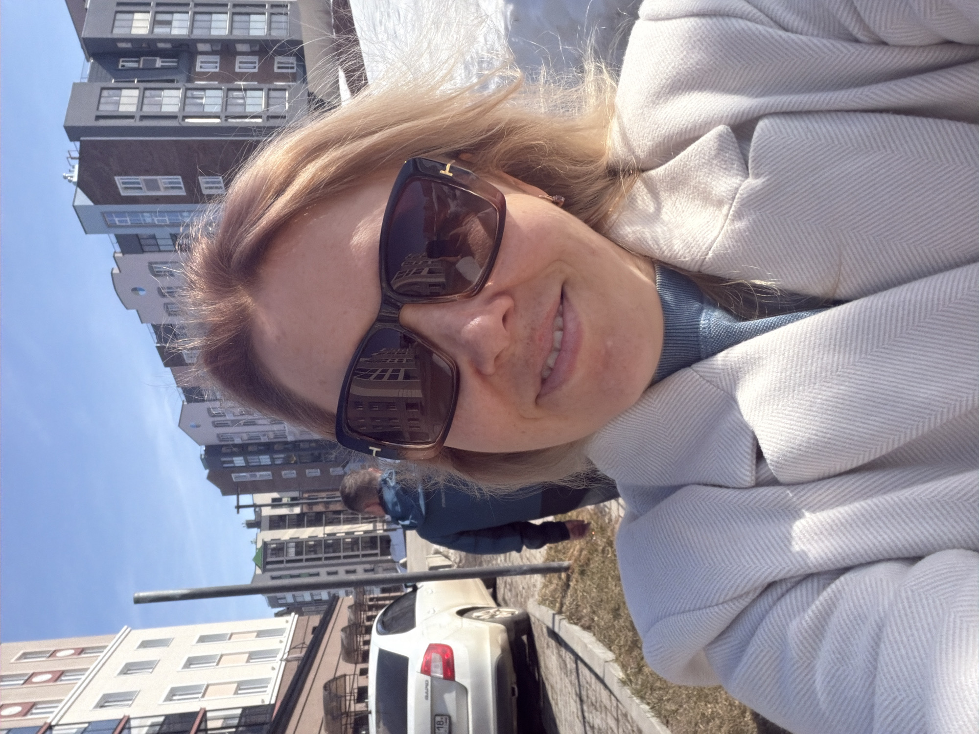 A woman is taking a selfie outdoors. She is wearing sunglasses and a light-colored coat. A city street scene is in the background. A white car is parked on the street. A man in a blue jacket is walking in the background. The building in the background has distinctive rectangular windows. The license plate on the white car is partially visible. The location is in Pervomaysky, Russia.