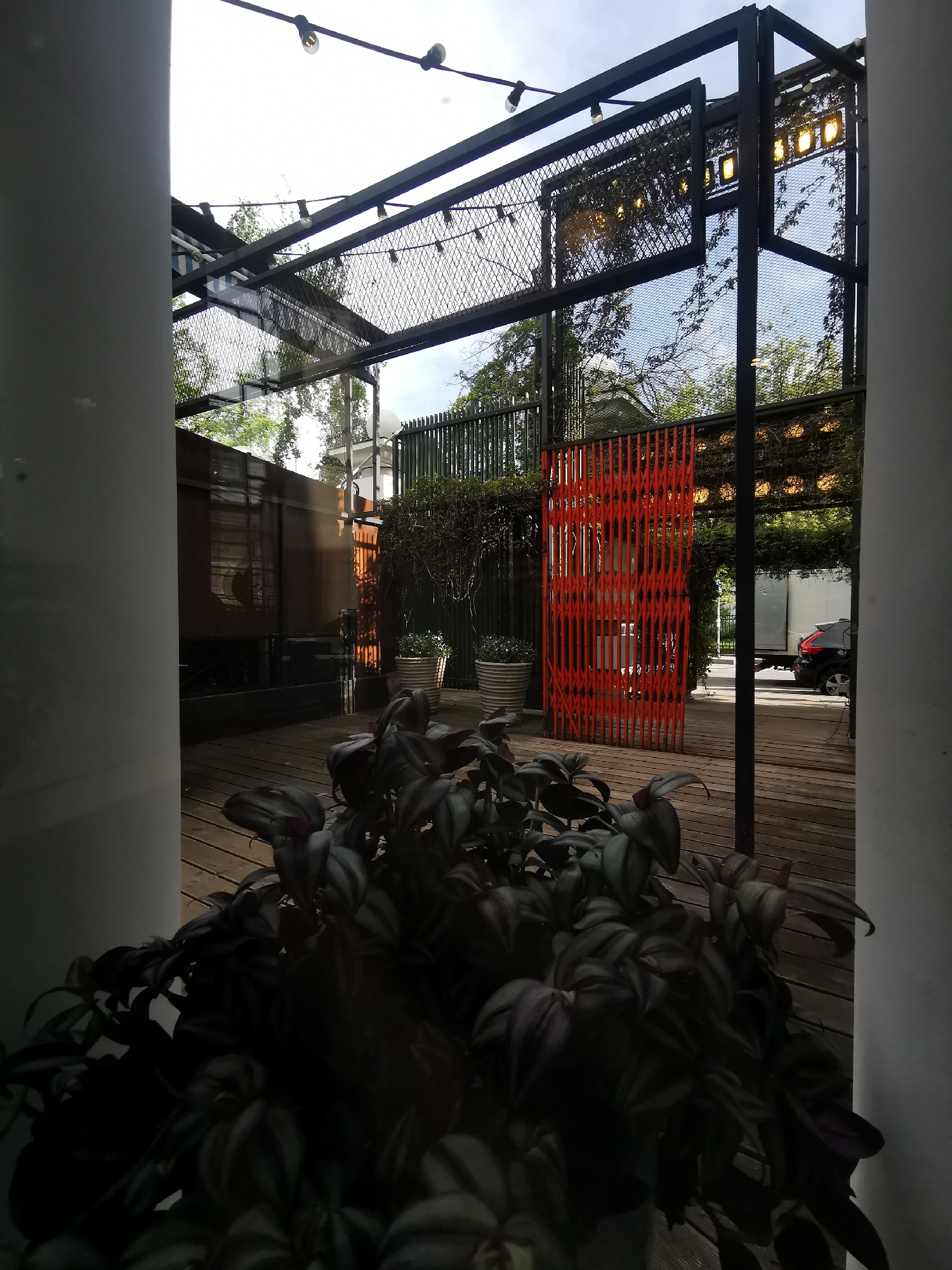 The image depicts an outdoor scene in Moscow, Russia, as suggested by the urban environment. The view is partially obstructed by a plant in the foreground, positioned between two white pillars. Beyond the pillars, there's a wooden deck area enclosed by a black metal structure with a mesh-like covering and string lights hanging above. An orange accordion-style security gate is visible, along with potted plants and a glimpse of a street with a car and a truck. The scene is captured during daylight, with a slightly overcast sky visible through the structure.