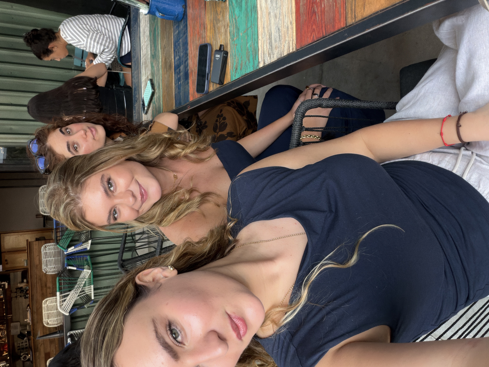 The image captures a group of four women sitting at a colorful, striped table. From the bottom of the frame upwards, the first woman is fair-skinned with blonde highlights and is wearing a dark blue top. Above her is another fair-skinned woman with long blonde hair, smiling at the camera. A third woman is partially visible, with brown curly hair and sunglasses on her head. Further in the background, a fourth woman in a striped shirt is looking at a phone, with a blue bag nearby. On the table, there is a phone and a small black electronic device. They are in a location with indoor seating with decorative chairs and wooden furniture visible in the background.