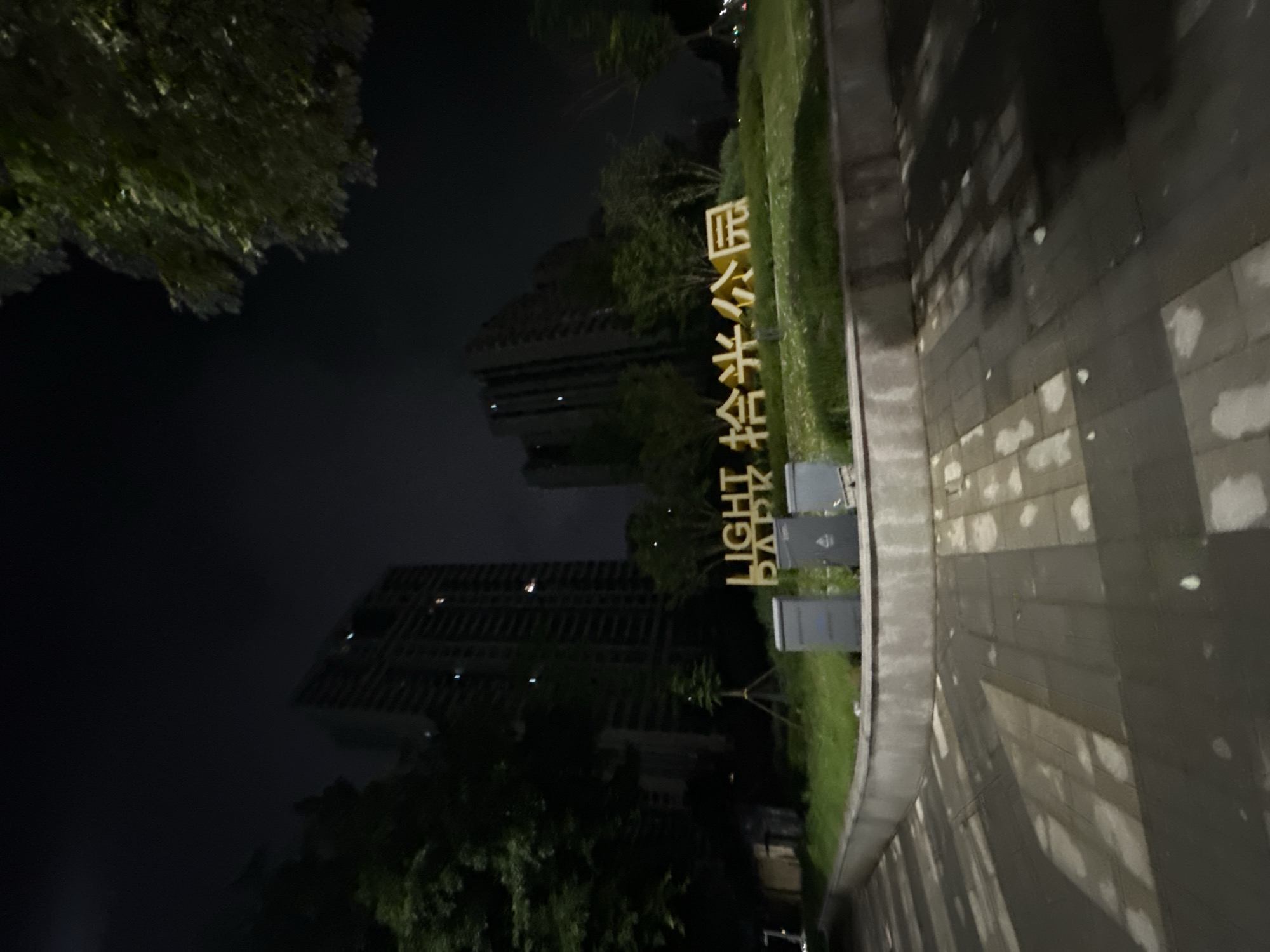 A low-angle, night-time exterior shot captures the entrance to "Light Park" in the Chongqing High-tech Zone, China. Two large, modern residential buildings with many illuminated windows rise in the background against a dark sky. In the foreground, a paved walkway with rectangular stones is partially illuminated by overhead lights, creating strong shadows. A low, curved concrete wall borders a grassy area where a large, illuminated sign in gold letters reads "LIGHT PARK" followed by Chinese characters. Three gray utility boxes are situated on the grass in front of the sign. Trees with dense foliage frame the scene on the left and right, with the right side of the image showing a glimpse of distant city lights.