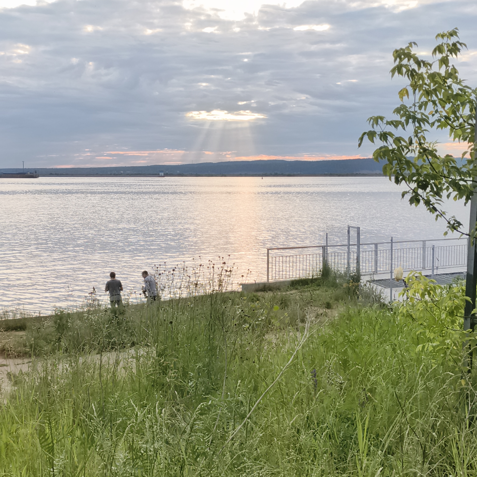 The image depicts a serene waterside scene in Chaikovsky, Russia, likely along the Kama River, given the city's prominent location on its banks. The foreground is dominated by lush, green grass and wildflowers, suggesting a natural embankment. Two individuals are visible standing near the water's edge. One, wearing a gray t-shirt and dark pants, faces away from the camera and towards the water, possibly fishing or observing the river. Beside him, another person, in a light-colored shirt and dark pants, also faces the water.
The water itself is calm, reflecting the overcast sky, with a notable sunbeam piercing through the clouds, creating a dramatic light effect over the water's surface. In the mid-ground, a metal railing and a paved walkway are present, indicating a developed area or pier. Further in the distance, the opposite bank of the river is visible as a low, tree-covered horizon line. A cargo ship or barge is also discernible on the left side of the horizon. The overall atmosphere is tranquil and hints at a late afternoon or early evening setting due to the soft, diffused light.