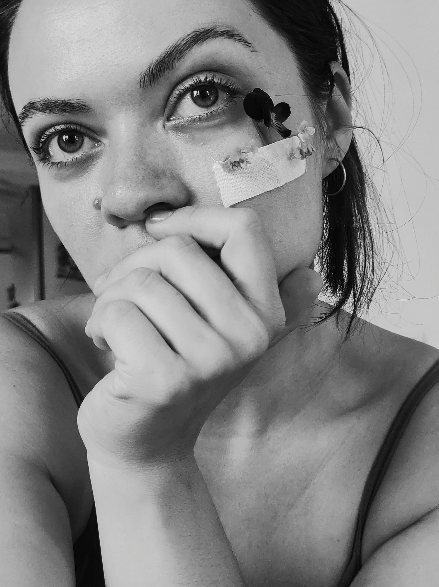 A black and white portrait shows a woman with dark hair pulled back, gazing intensely at the camera. Her hand is raised to her mouth, with her fingers resting on her lips. A bandage is adhered to her cheek near her eye, decorated with small white flowers and a single, dark flower. The location is Unknown, Unknown.