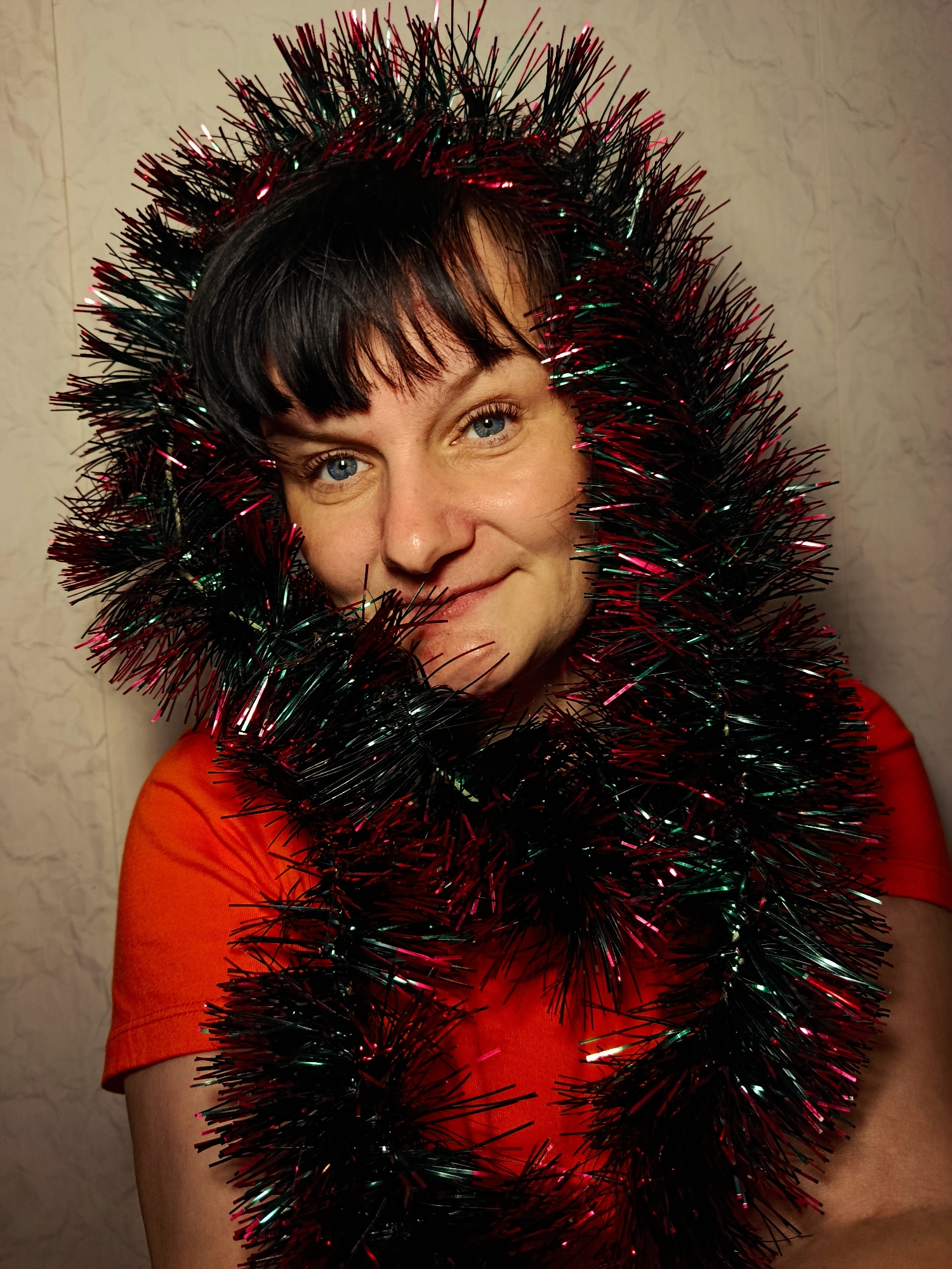 A person with short, dark hair and blue eyes is wearing a red T-shirt and is framed by a thick, multi-colored tinsel garland. The garland is a mix of black, green, and red strands, appearing to create a halo effect around the person's head and shoulders. The background is a light, textured wall. The person is looking at the camera with a slight smile. The location in the city of Фоки, Russia, cannot be determined from the image.