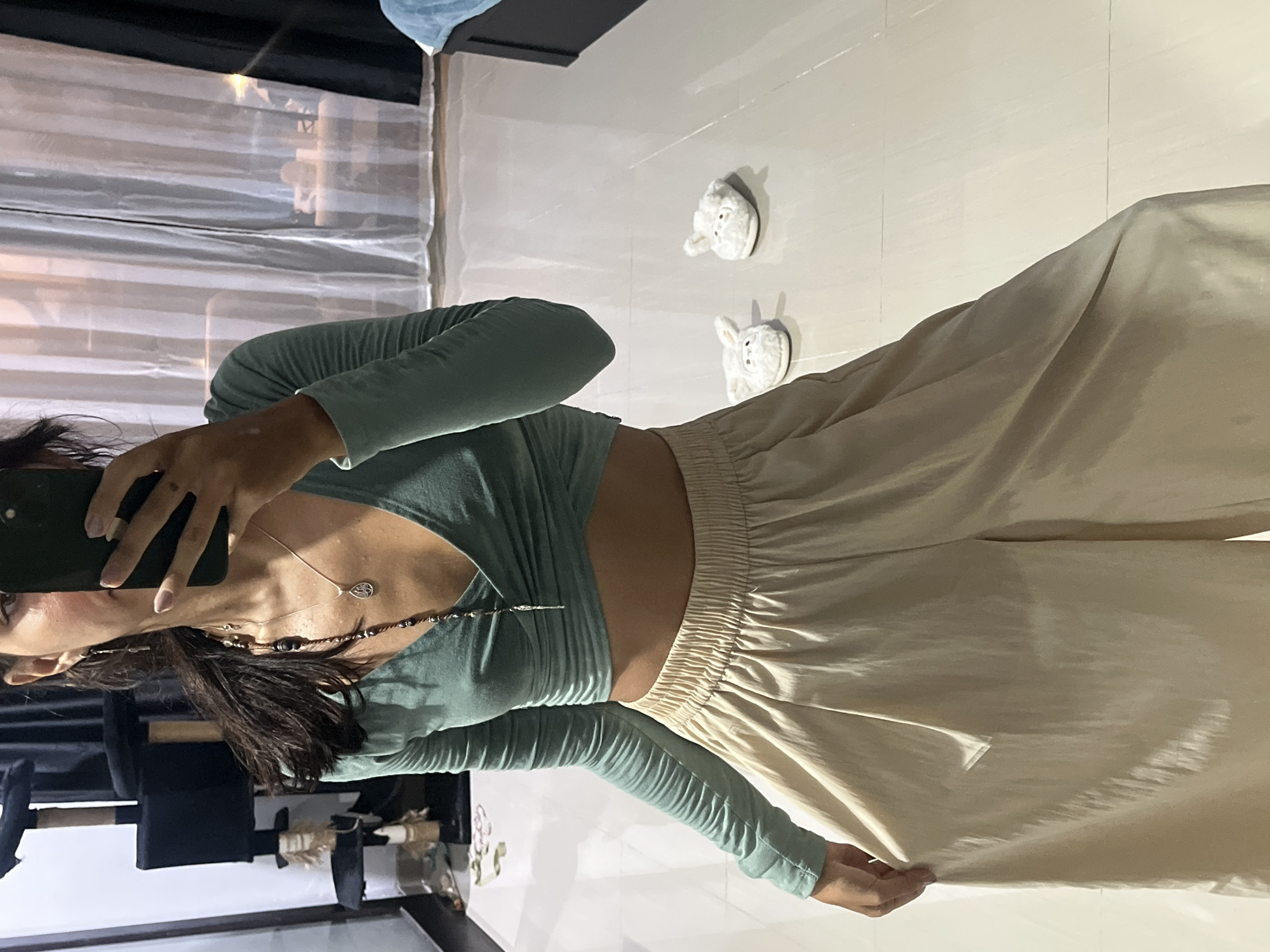 A person, likely a woman, is captured in a vertical, waist-up mirror selfie, showcasing her outfit. She holds a black smartphone to take the picture, partially obscuring her face and extending across her left cheek.
She wears a long-sleeved, light teal or mint green cropped top with a crossover design at the front, revealing a strip of her midriff. Her bottom garment consists of light beige or cream-colored wide-leg pants with an elasticated, ruched waistband. She has long, dark hair flowing over her shoulders. Two necklaces are visible: one with a small silver leaf-shaped pendant and another layered, beaded necklace. Her fingernails are painted a light, neutral color.
The setting is indoors, featuring a light-colored tiled floor. Two white, fluffy slippers, each designed with an animal face (resembling a rabbit), are placed on the floor behind her. The background behind the person is dark, possibly a piece of furniture, and reflects a light-colored, horizontally striped surface, suggesting a window with blinds or a decorative panel. Other blurry objects can be seen on a surface in the lower left.
The image has a casual, domestic feel, likely taken in a home or private space in Kuta Selatan, Indonesia. The lighting is soft and even, consistent with indoor conditions. No visible text is present in the image.