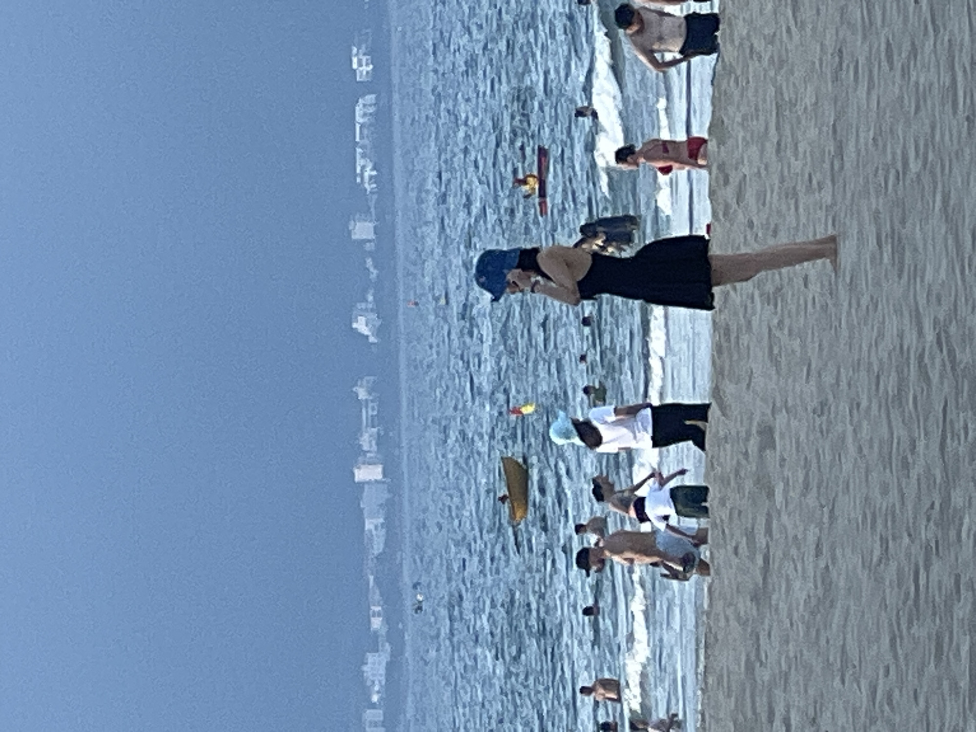 A medium shot captures a crowded beach scene in Đà Nẵng, Vietnam, with the ocean stretching to a hazy horizon lined with distant buildings. In the foreground, a woman in a black dress and blue hat stands on the sand, facing left. Behind her, the water teems with people swimming and wading. A yellow boat is visible further out in the sea, and a red boat with a person in it is to the right. The sky is a muted blue, suggesting overcast conditions or dusk. The waves are gentle, lapping at the shore where various individuals, including children, are enjoying the water. The overall impression is one of a lively beach day in a coastal Vietnamese city.