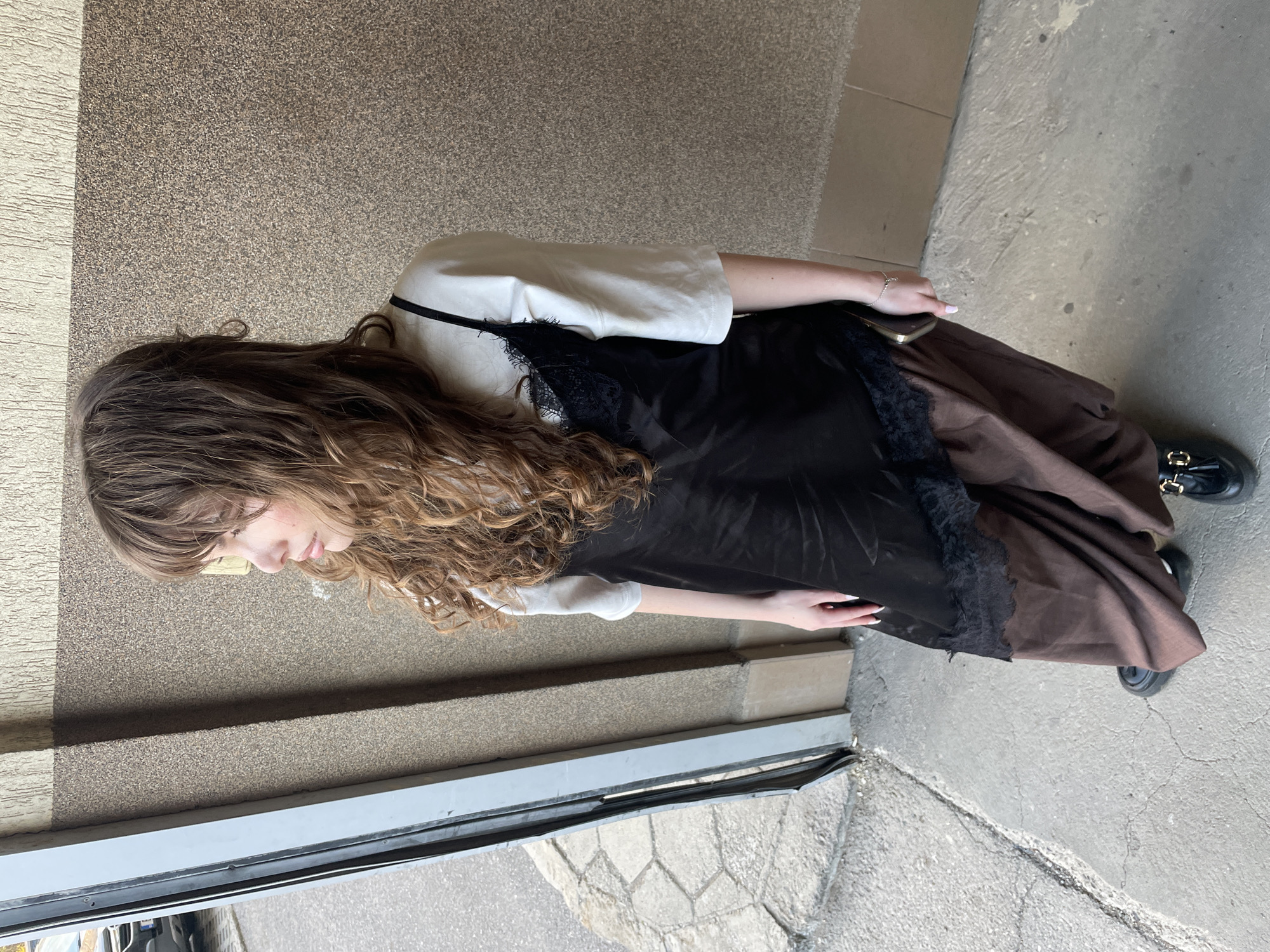 A young woman stands in an urban environment, likely in Sofia, Bulgaria given the request context. She is wearing a white short-sleeved t-shirt underneath a black slip dress with lace detailing, and a long brown skirt. She has long, curly brown hair. She is holding a phone in one hand and her fingers are painted with light nail polish. She is wearing black shoes with a gold-colored decoration. The scene appears to be outdoors, adjacent to a wall with a rough texture and a tiled floor. A door with a silver frame can be seen.