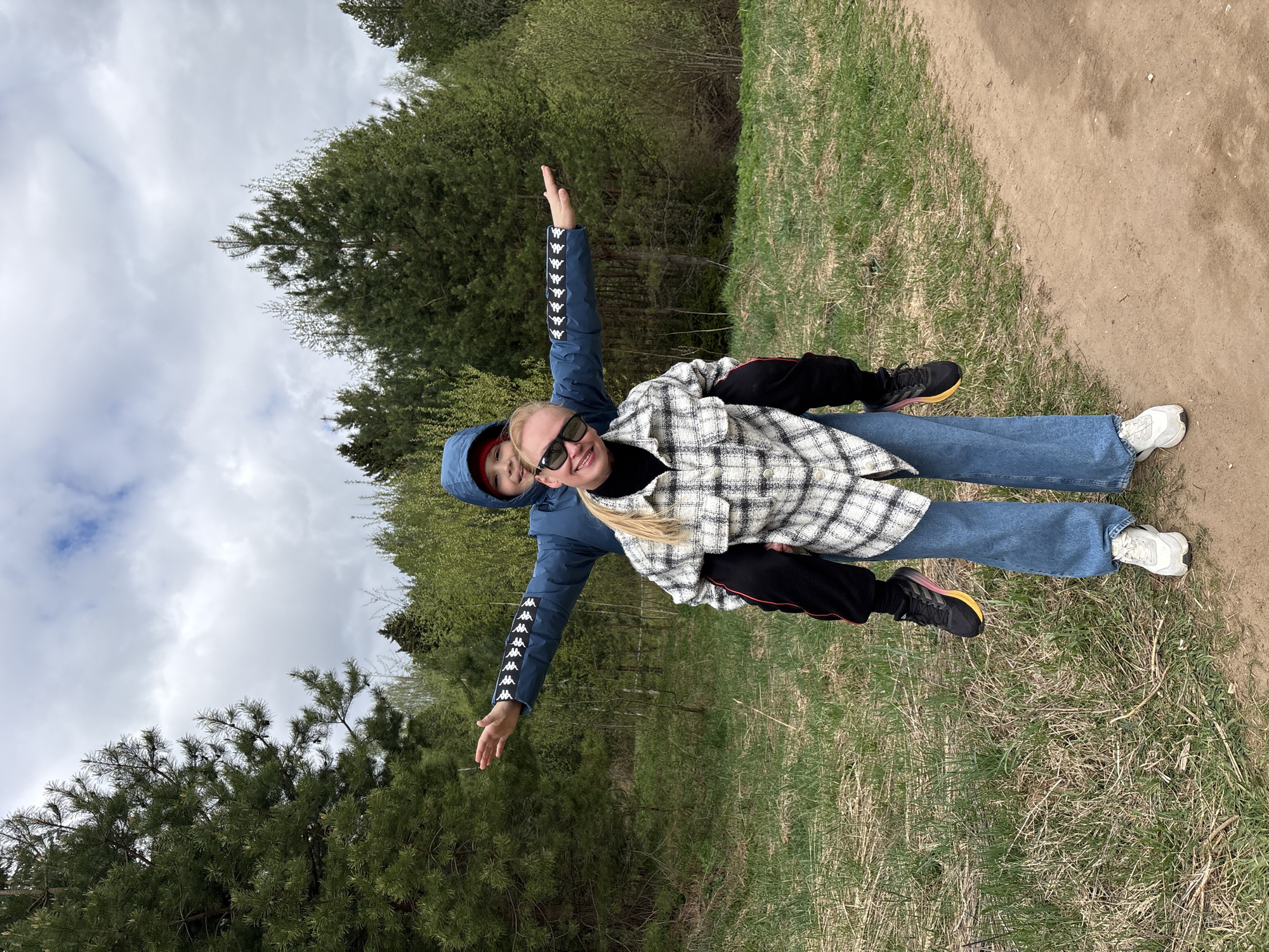 Two people are standing on a dirt path with grass on either side. A woman wearing sunglasses, a plaid jacket, and blue jeans is standing in front, while a child wearing a blue hooded jacket is standing behind her. Both have their arms outstretched. A forest is visible in the background. The location is Chaikovsky, Russia.
