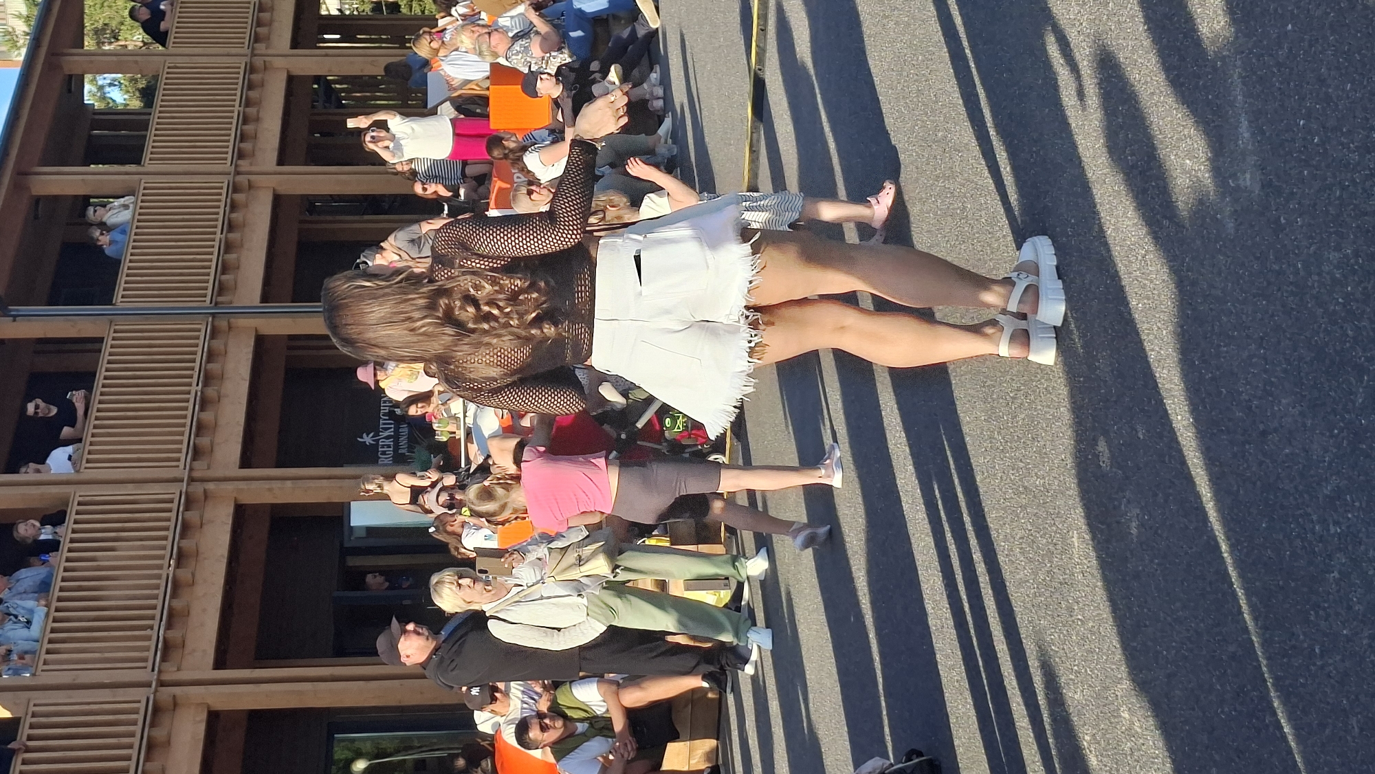 In Tallinn, Estonia, on a paved outdoor area, a group of people are gathered. In the foreground, a woman with long, wavy brown hair is dancing, wearing a black mesh top and white frayed shorts with chunky white platform sandals. To her left, a young girl in a pink crop top and grey cycling shorts dances, and a woman in a white cardigan and green pants observes. In the background, other individuals are seated or standing, some on a wooden deck with railings, suggesting a venue with multiple levels. The setting appears to be a sunny daytime event, with shadows cast across the ground by structures and people.