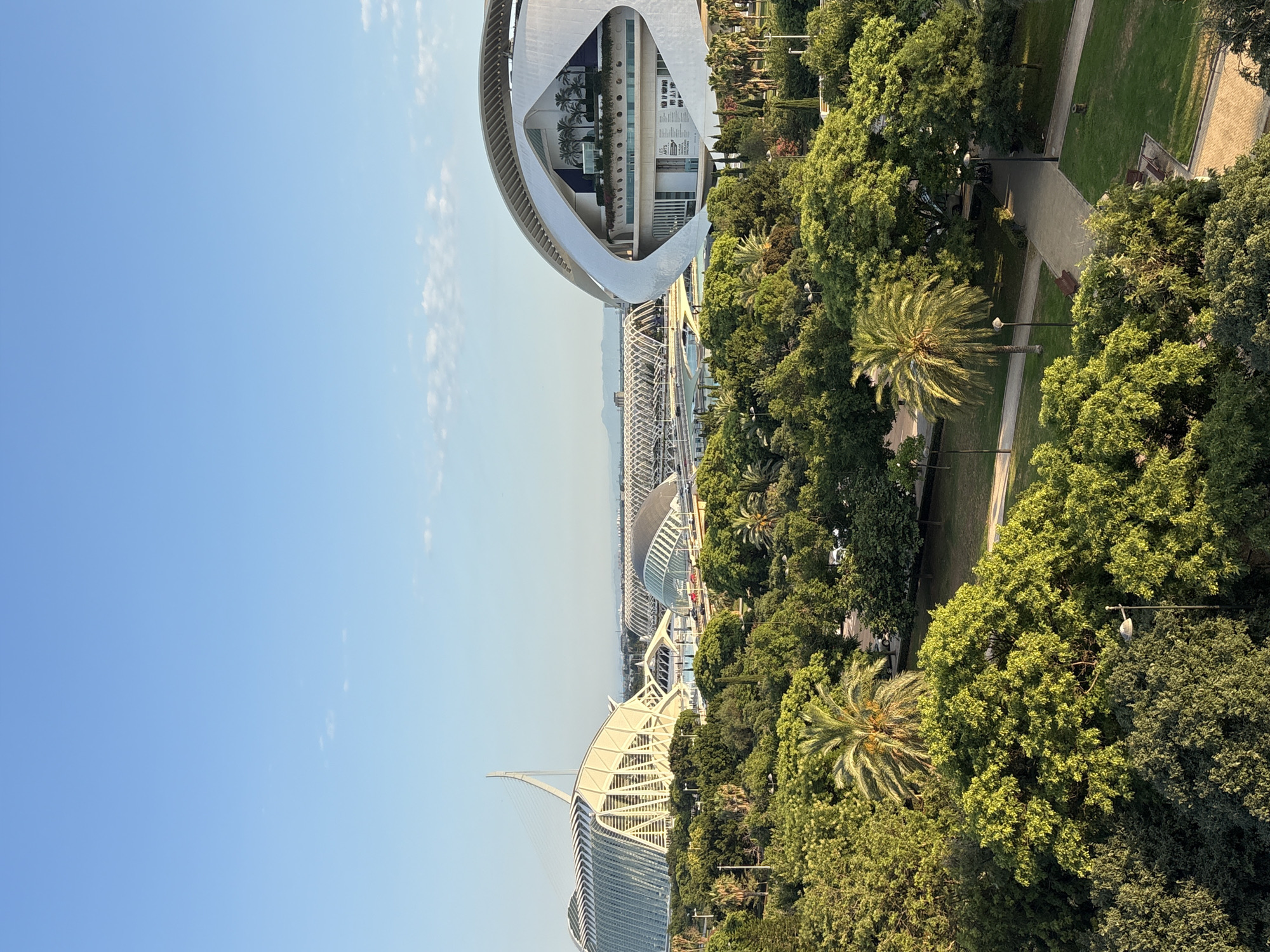 The image depicts an aerial view of the City of Arts and Sciences in València, Spain. The iconic architecture of the complex is visible, including the distinctive curved white structure of the Hemisfèric on the left, and the ribbed white structure of the Palau de les Arts Reina Sofía in the foreground. These modern buildings are surrounded by lush green trees, palm trees, and manicured park areas with pathways and benches. The scene appears to be a bright, sunny day, with a clear blue sky and scattered white clouds. In the distance, more of the complex's futuristic buildings and structures can be seen, along with hints of the urban landscape and mountains beyond. The overall impression is one of a serene and well-maintained public space, bathed in warm sunlight, suggesting it's likely late afternoon or early evening due to the quality of the light and shadows. No people are prominently visible, but the presence of benches and pathways indicates it is a space designed for public enjoyment. The image is oriented vertically.