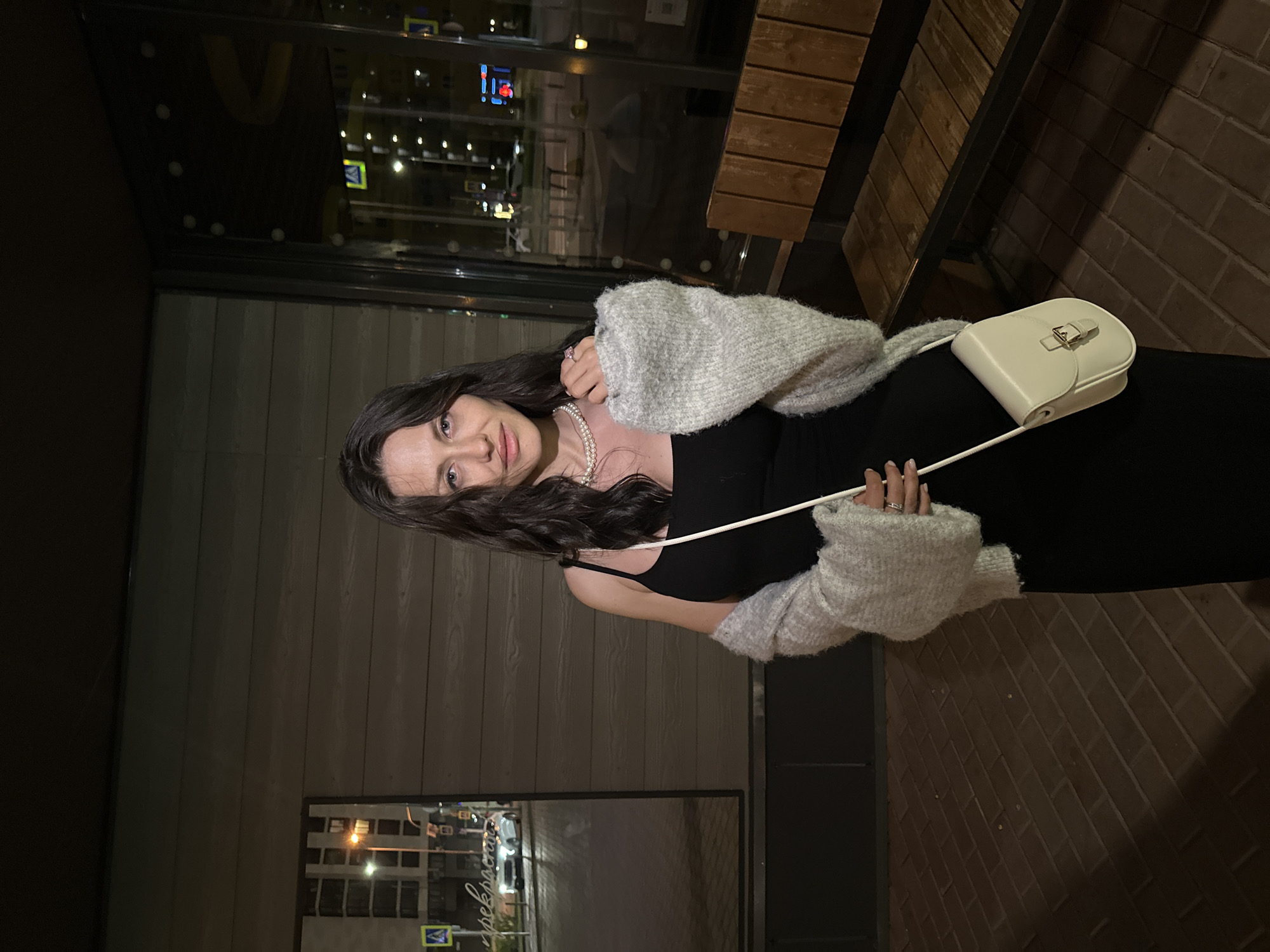 A full-shot image taken at night depicts an adult female standing outdoors in front of a building with horizontally paneled dark brown siding. The woman, who appears to be in her 20s or 30s, has long, dark, wavy hair and is looking directly at the camera with a neutral expression. She wears a black sleeveless midi-dress and a light grey, loose-knit cardigan draped off her shoulders. A double-strand pearl necklace adorns her neck, and a silver ring is visible on her right hand. She carries a small, cream-colored crossbody purse with a buckled flap closure, held by a thin strap over her left side. The ground beneath her is paved with reddish-brown bricks in a stretcher bond pattern.
To the left of the woman, a large reflective surface (likely a window or mirror) reflects a street scene, including a white car, streetlights, a multi-story building, and a blue pedestrian crossing sign. Faint Cyrillic text, "прекрасна", is visible on or through this surface. To the right of the woman, a large glass window reflects distant city lights, blurred vehicles, and illuminated signs (including red and blue lights), while revealing an interior with a yellow architectural feature. A wooden bench with a dark metal frame and horizontal wooden slats is positioned against the building wall to the right. The scene is illuminated by various artificial light sources typical of an urban environment at night. The specific city and precise location are unknown, but the setting is clearly an urban street adjacent to a commercial or residential building.
