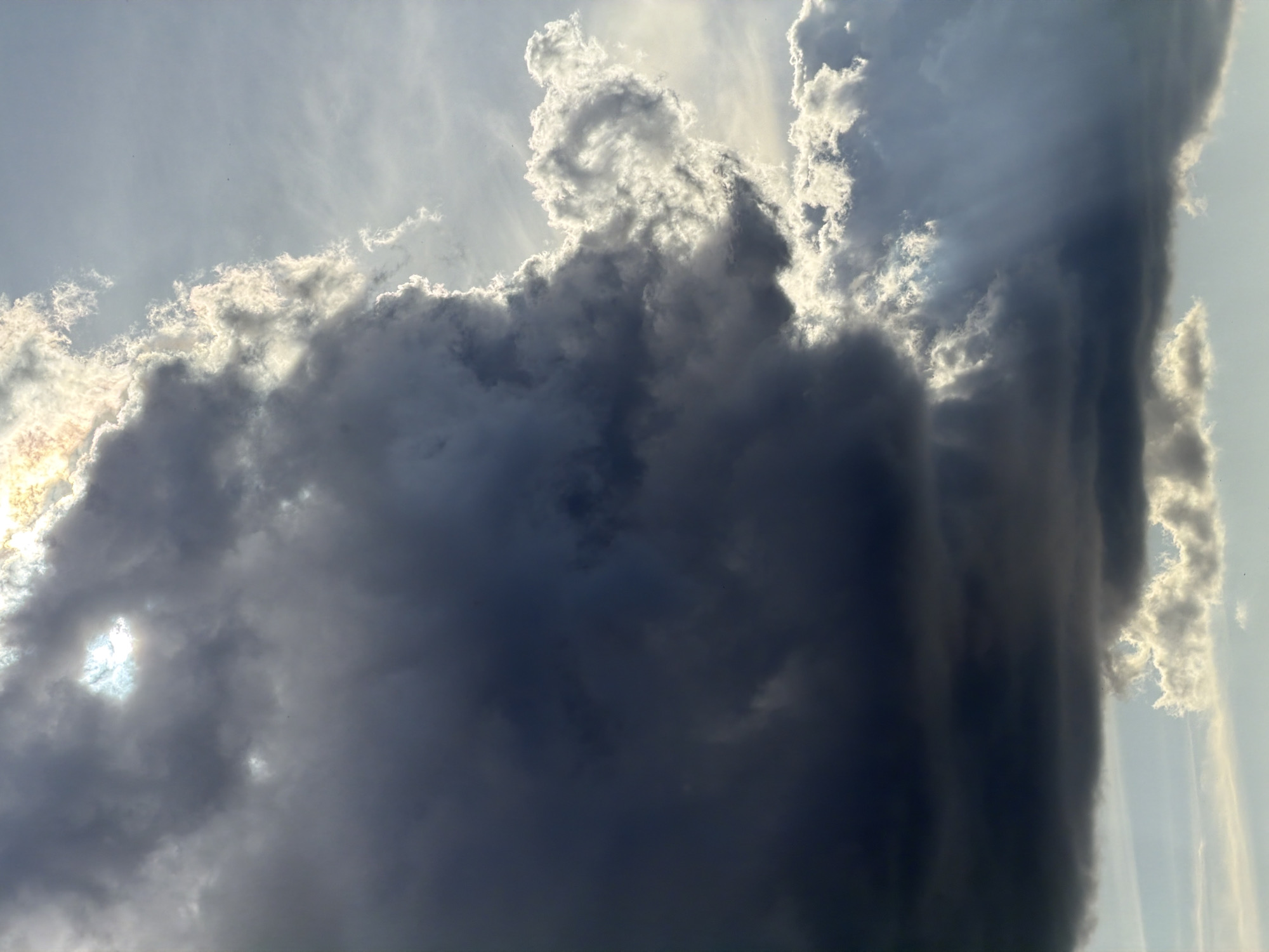 The image depicts a sky filled with dark, heavy storm clouds. The clouds are a mix of gray and dark blue, with some edges illuminated by sunlight. The sunlight seems to be breaking through some parts of the cloud cover, creating areas of bright light against the dark clouds. There are no people, subjects, or actions visible in the sky.
The environment is the sky during a thunderstorm with sunrays visible.
The location in Krasnoyarsk, Russia, is not identifiable from the image.