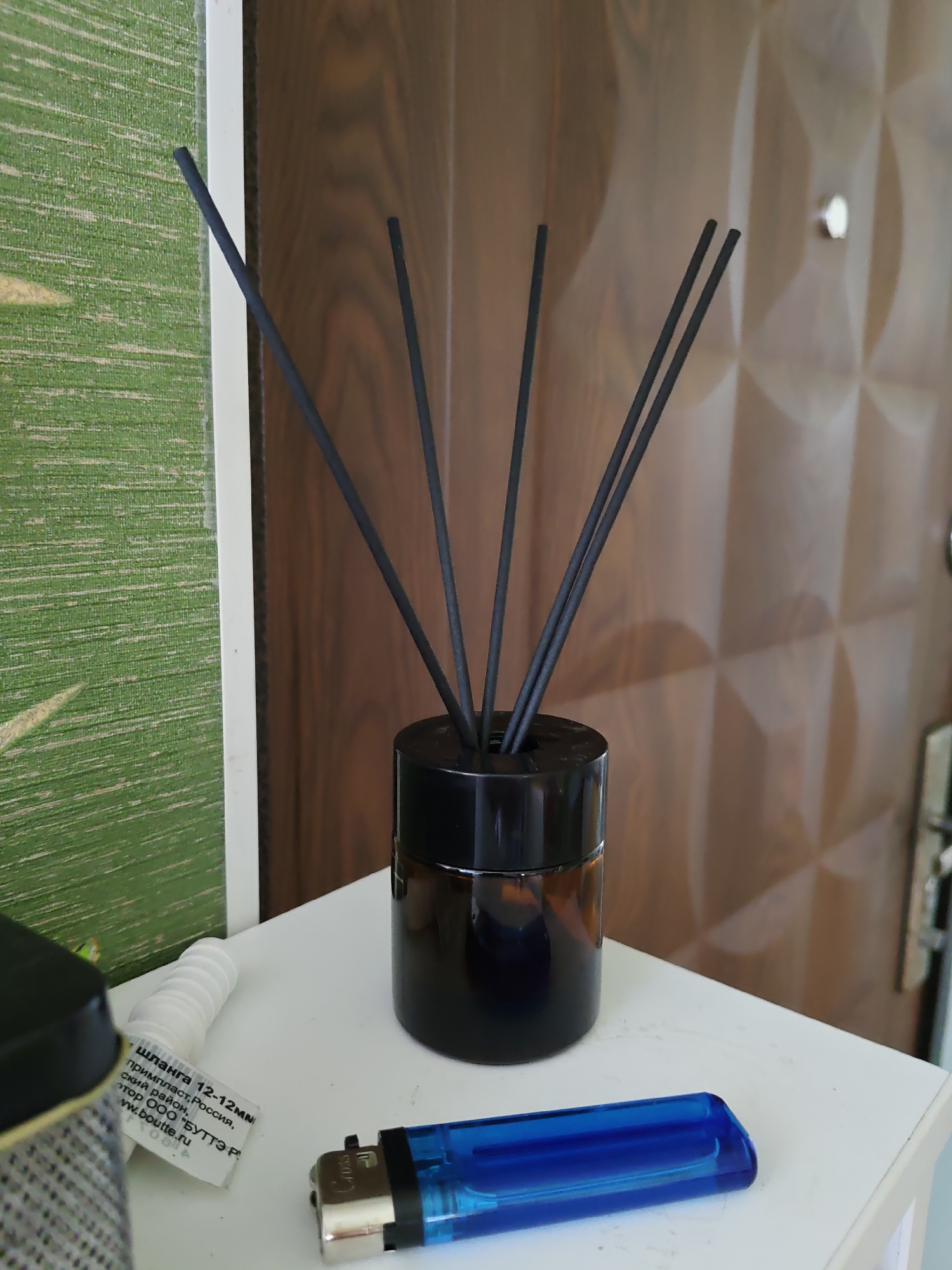 A dark brown glass reed diffuser bottle with black reeds stands on a white surface in front of a wood-paneled wall. To the left of the diffuser is a blue and silver lighter, and next to that is a white coiled object with a label that appears to be in Russian. The background wall has a distinctive geometric pattern of overlapping circles. The overall scene suggests an indoor setting, possibly a home or office, with a focus on decorative and everyday objects. There is no indication of the specific location within Rostov-on-Don, Russia.