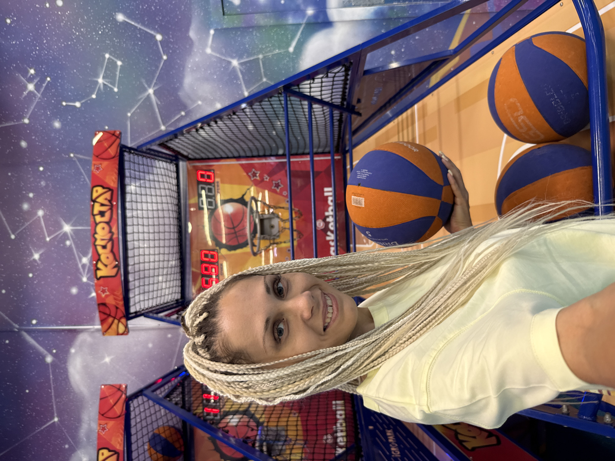 A woman with long blonde braids is standing in front of a basketball arcade game. She is wearing a light yellow t-shirt and is holding a blue and orange basketball. The arcade game has a digital score display showing "11" and "9.88". Behind the game, a mural depicts a starry sky with constellations. The flooring is orange. The image appears to be taken indoors, likely at an entertainment center or arcade in the city of Rusanovo, Russia.
