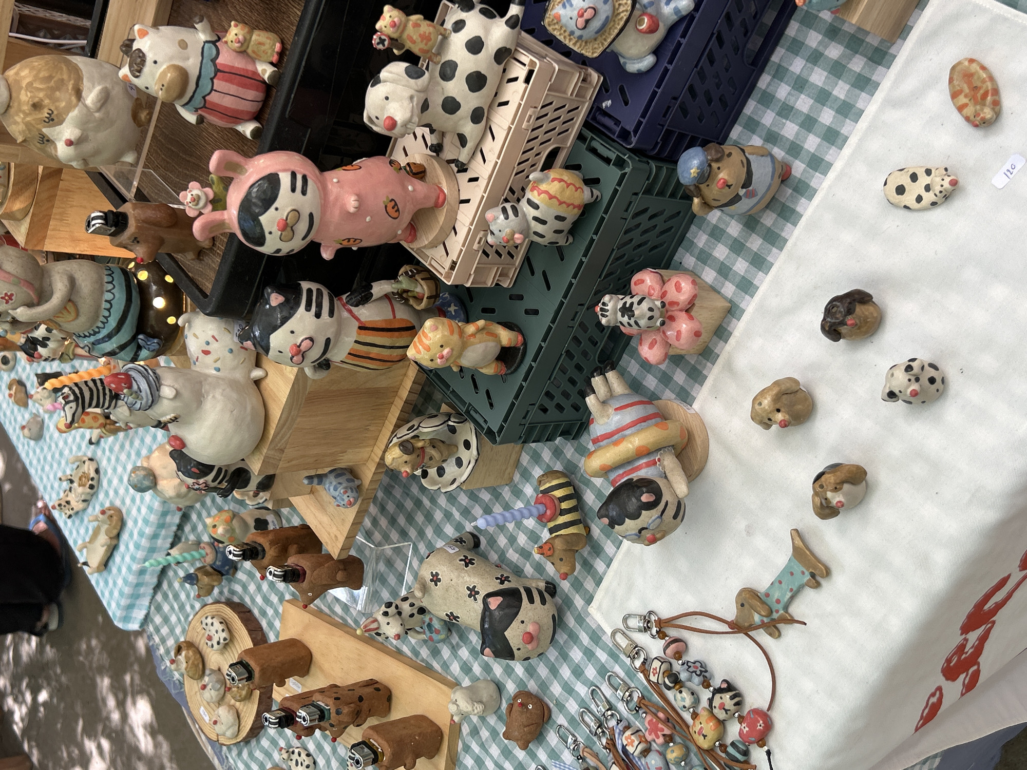 A market stall in Ban Sai Kham, Thailand, displays an assortment of handmade ceramic animal figurines. The figurines, primarily dogs and cats, are rendered in a whimsical, stylized manner with various patterns and colors, including polka dots, stripes, and floral motifs. Some are posed playfully, while others appear to be functional objects like lighters or incense holders. The stall is covered with a green and white checkered tablecloth, and the items are arranged on wooden shelves, crates, and directly on the table. In the background, a person's legs are visible, suggesting a vendor or shopper. The overall scene conveys a creative and artisanal atmosphere.