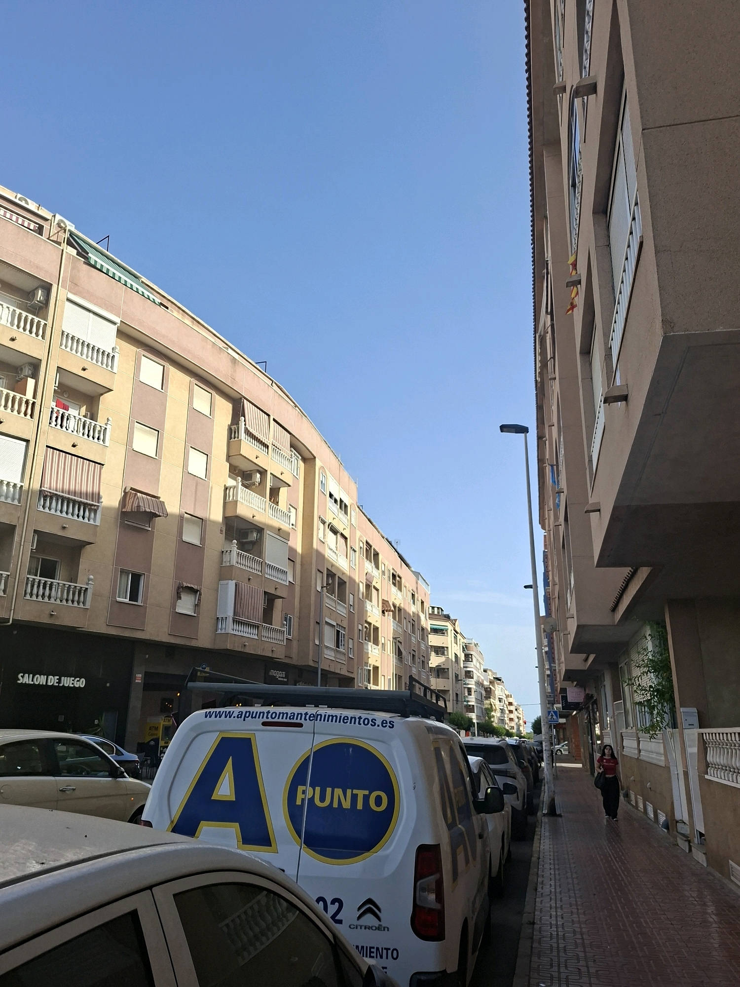 This is a street scene in Torrevieja, Spain, characterized by apartment buildings lining a pedestrian walkway. The buildings on the left are painted in shades of pink and beige with multiple balconies, while the building on the right is a solid beige structure. A white van with large blue and yellow lettering, "A PUNTO," is parked on the street, partially obscuring other vehicles. The van has the website "www.apuntomantenimientos.es" printed on its side. A woman in a red shirt and dark pants is walking on the right-hand sidewalk. The sky is clear and blue, indicating a sunny day. A street lamp is visible on the right side of the image.