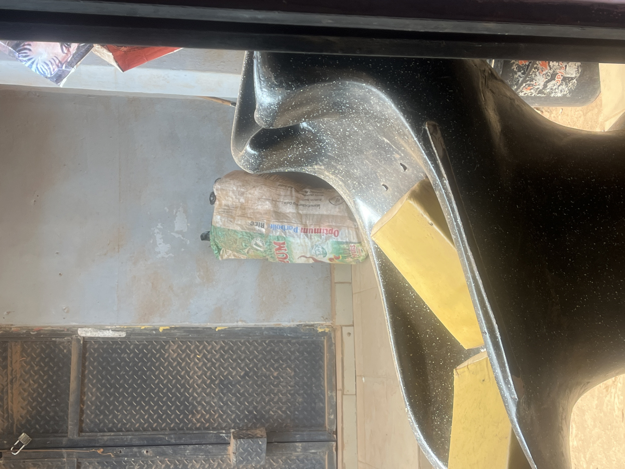 A bag of Optimum Parboiled Rice is visible, sitting on what appears to be a metal floor panel with a diamond plate pattern. The bag is positioned near a wall and next to what looks like a black, possibly plastic, object with yellow components. The location is likely somewhere in Abuja, Nigeria, given the product brand's distribution. The scene is viewed from an elevated perspective, potentially through a window. No people are visible.