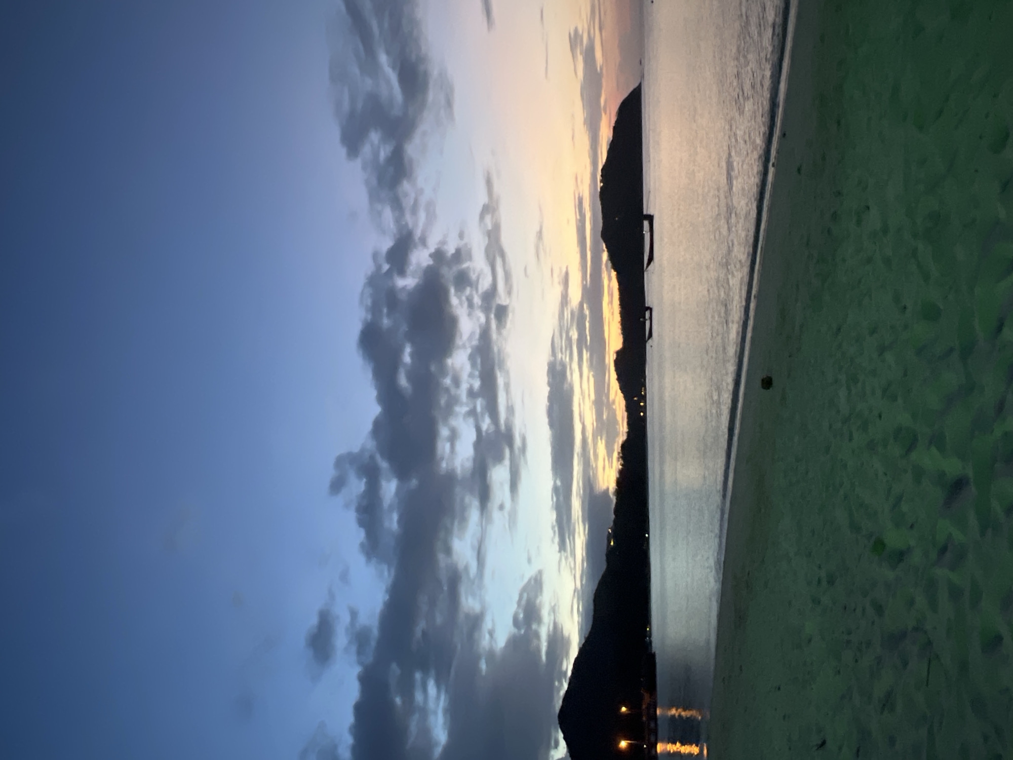 The image depicts a serene beach scene at dusk in Ko Pha-ngan District, Thailand. The sky displays a gradient of deep blue transitioning to pale orange and pink near the horizon, with scattered dark clouds. Silhouetted against the sky are two islands, one to the left with lights visible from what appears to be a pier or waterfront structures, and another larger island to the right. The calm sea reflects the sky's colors. Two small boats are visible on the water. The foreground is a sandy beach, with the sand appearing a muted green in the low light. A single coconut is present on the sand. The overall impression is one of peaceful twilight on a tropical coast.