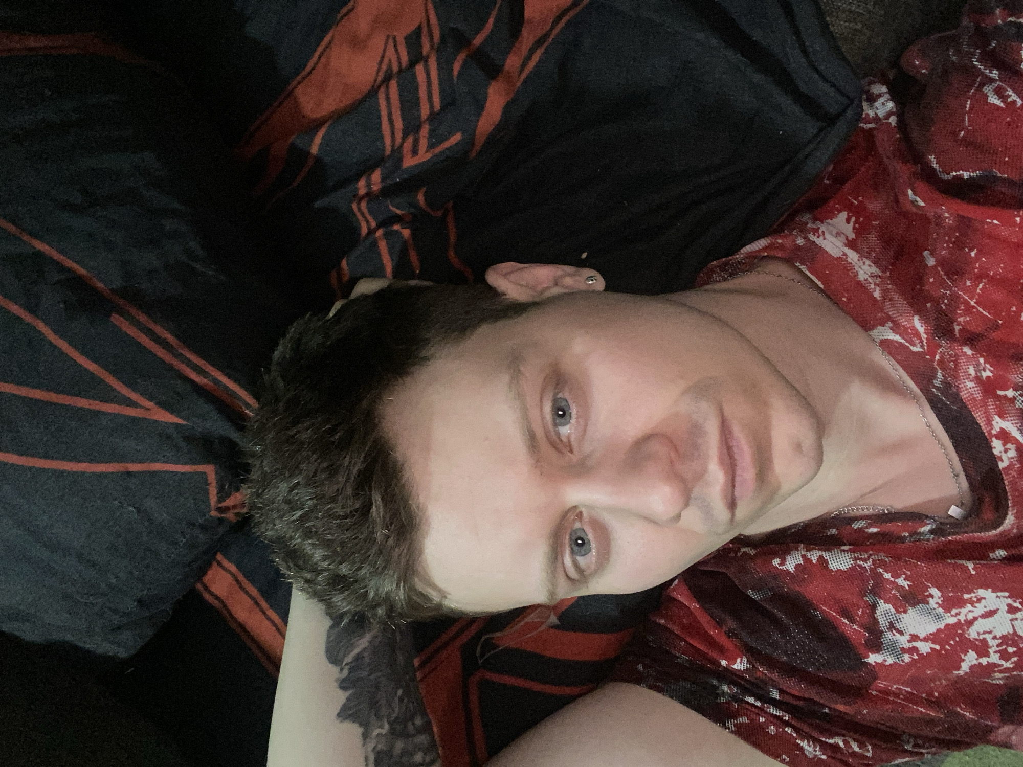 The image is a close-up selfie of a person with fair skin and blue eyes, lying down on what appears to be a bed with dark and red striped bedding. The person has short, styled brown hair and is wearing a red t-shirt with a distressed pattern. They have a tattoo on their left arm, which is partially visible, and a silver earring in their left ear. The person is looking directly at the camera with a neutral expression. The lighting is dim, casting shadows on the face and surroundings. There is no visible indication of the specific location within the city of Samara, Russia.