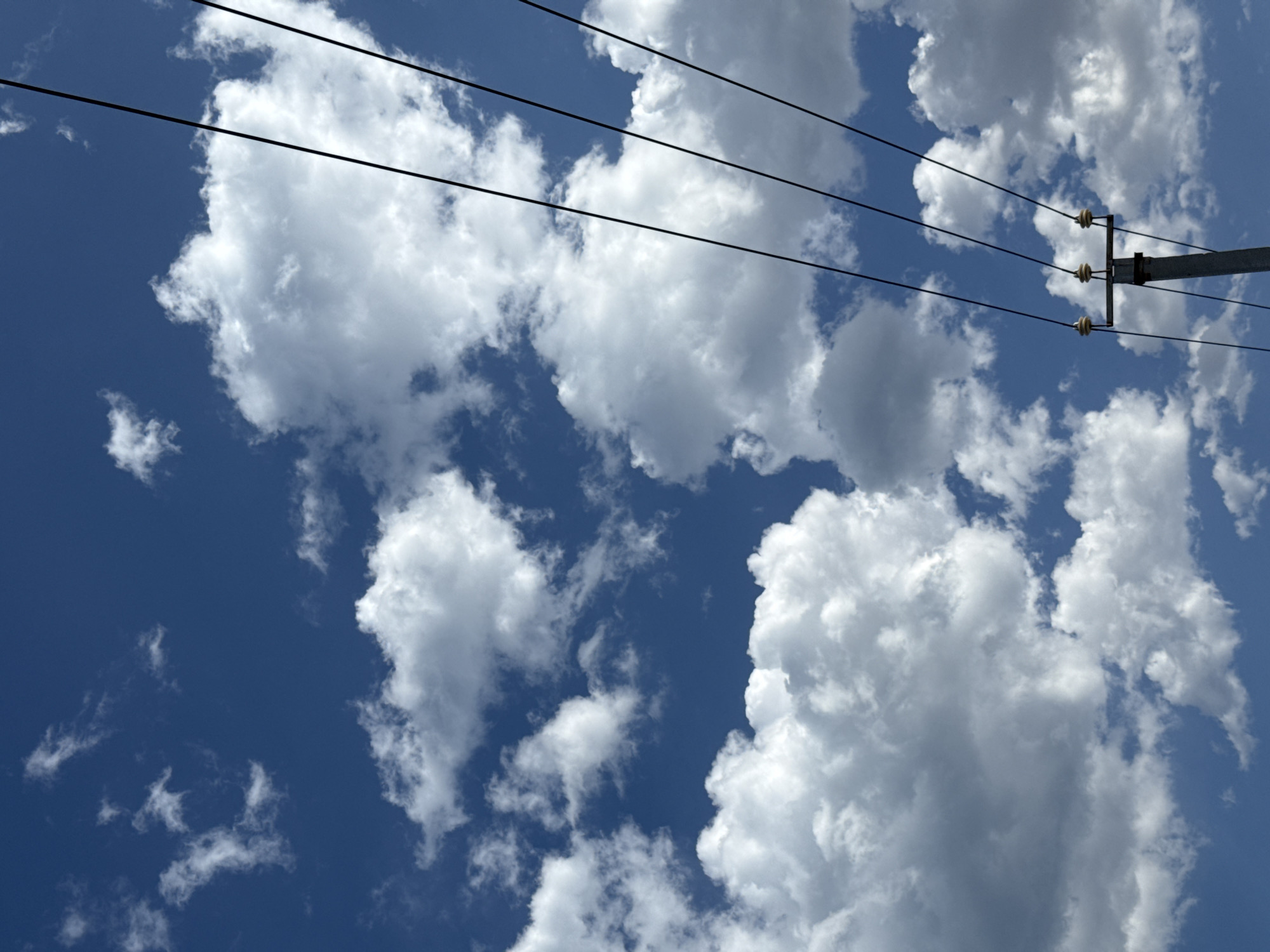 The image depicts a bright, cloudy sky with a clear blue expanse. The clouds are fluffy and white, with some casting shadows. In the foreground, a power pole is visible, supporting four black electrical wires that stretch diagonally across the frame from right to left. The pole itself is gray and metallic, with three ceramic insulators at the top where the wires are attached. The scene appears to be outdoors in a location that is possibly rural or suburban given the presence of the power pole. The overall impression is one of a clear day with scattered clouds. The location is identified as Ақши, Kazakhstan.