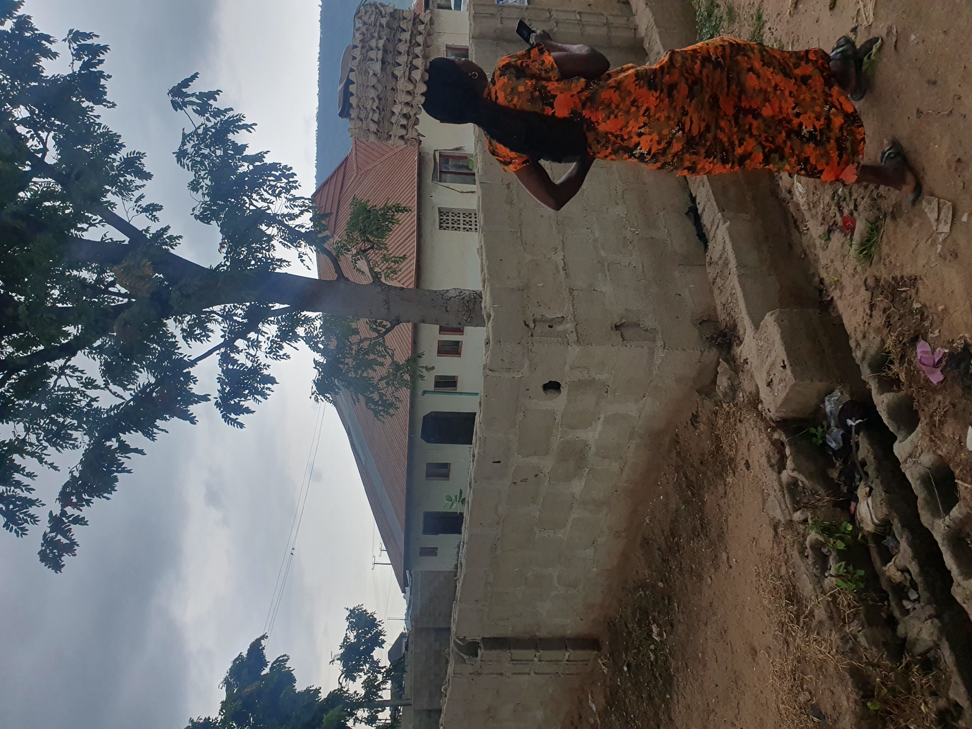 The image shows a woman walking in a dusty, open area in Lokoja, Nigeria. She is wearing a long, orange and black patterned dress and is holding a smartphone in her right hand. Her hair is long and black, and she has earrings on. She is walking away from the camera, with her left hand on her hip. Behind her is a beige brick wall and a house with a red-tiled roof. There are trees and some debris on the ground, suggesting an outdoor, possibly rural or semi-urban environment. The sky is overcast. The woman is walking past a drainage ditch filled with debris and some small plants.