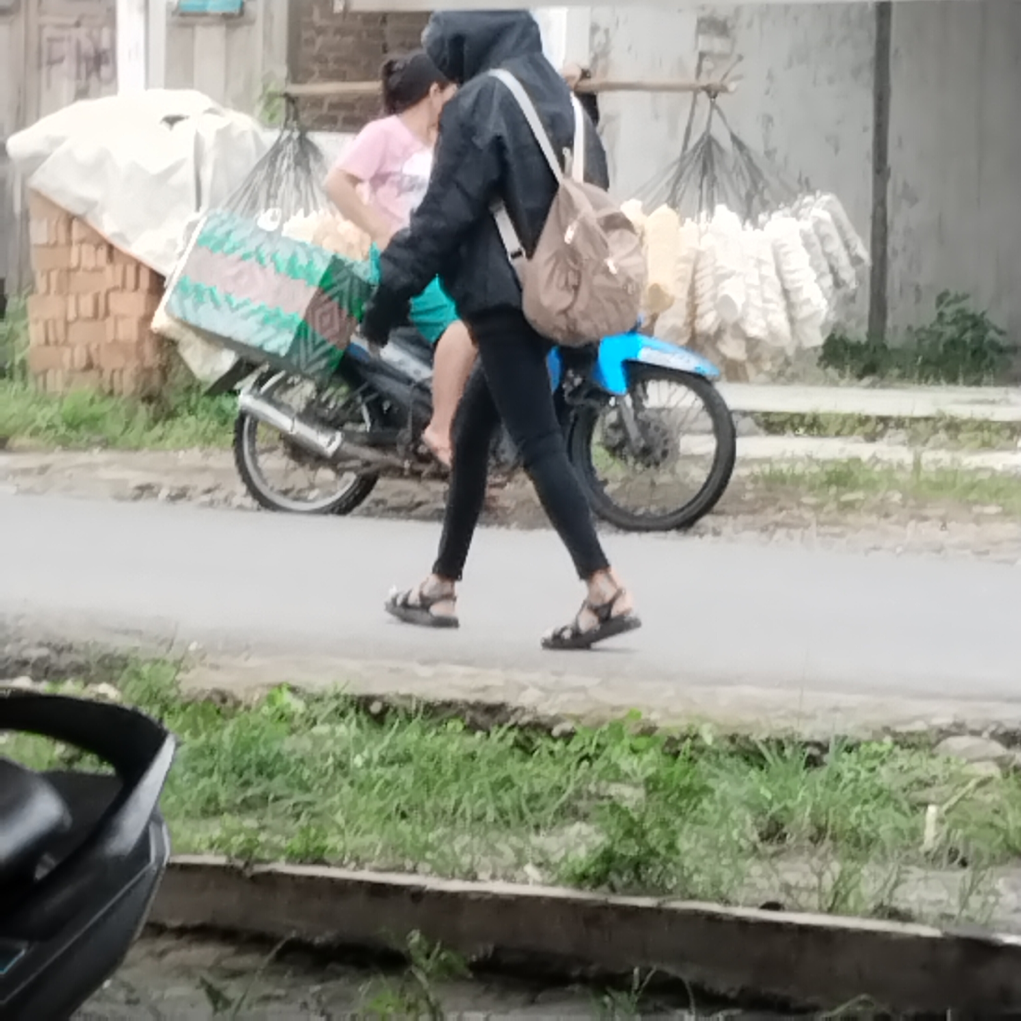 The image depicts a street scene in Talun Kenas, Indonesia. Two individuals are present. One, a person wearing a black hooded jacket and black pants, is walking with a brown backpack. This individual is wearing black sandals. Another person, wearing a pink t-shirt and dark shorts, is seated on a motorcycle. The motorcycle has a blue body and a large, patterned box strapped to its rear. The background shows a building and a structure from which items are hanging, possibly dried goods. The ground is paved with asphalt, and there is vegetation along the side of the road. The scene appears to be during daylight hours.