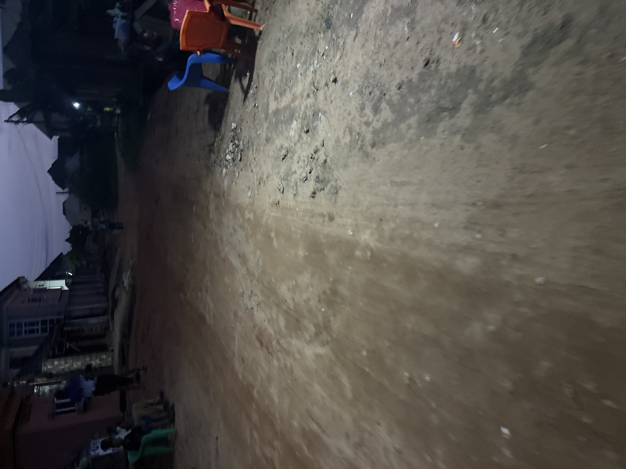 The image captures a dirt road in Obosi, Nigeria, at dusk. Several plastic chairs, including a blue one and orange and red ones, are arranged near a building on the right side of the frame. In the background, residential buildings are visible, some appearing to be under construction. On the left side, a few individuals are seated on a green plastic chair. The lighting suggests it is late evening, with ambient light casting shadows and creating a dim atmosphere. The overall scene depicts a common street view in a Nigerian residential area.