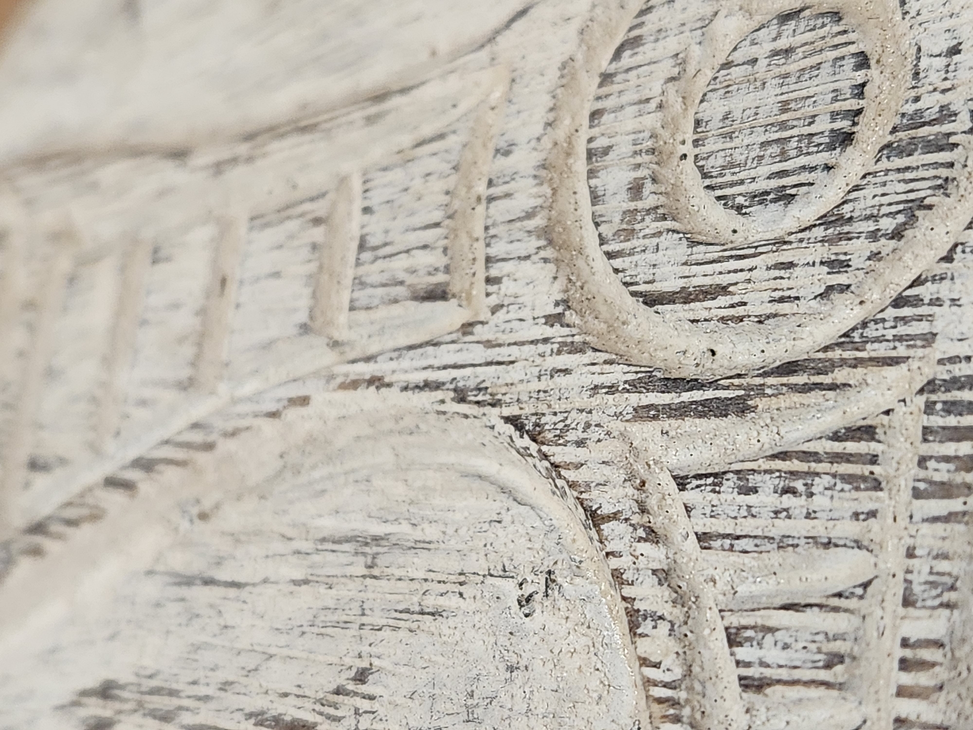 The image is a close-up, macro photograph of a distressed, carved wooden surface. The wood has a visible grain with vertical lines and is painted with a weathered, off-white finish that is chipped and worn, revealing darker wood underneath. There are carved details in the wood, including circular and linear patterns. The lighting creates shadows that emphasize the texture and depth of the carvings. The scene appears to be an object, possibly furniture or an architectural element, with a textured and aged appearance. The location is identified as Maitland, United States of America.