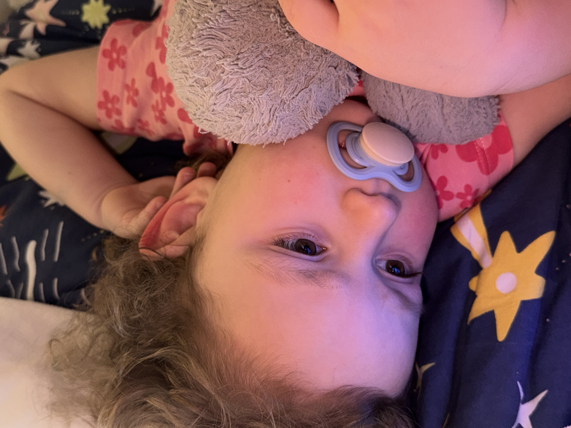 A young child is lying on their back on a bed with a dark blue comforter featuring yellow star patterns. The child has light skin, brown eyes, and curly blonde hair. They are wearing a pink shirt with a floral pattern and holding a gray stuffed animal. The child has a blue and peach-colored pacifier in their mouth. The photograph appears to have been taken indoors. The location in the city of Trassem, Germany cannot be determined from the image.