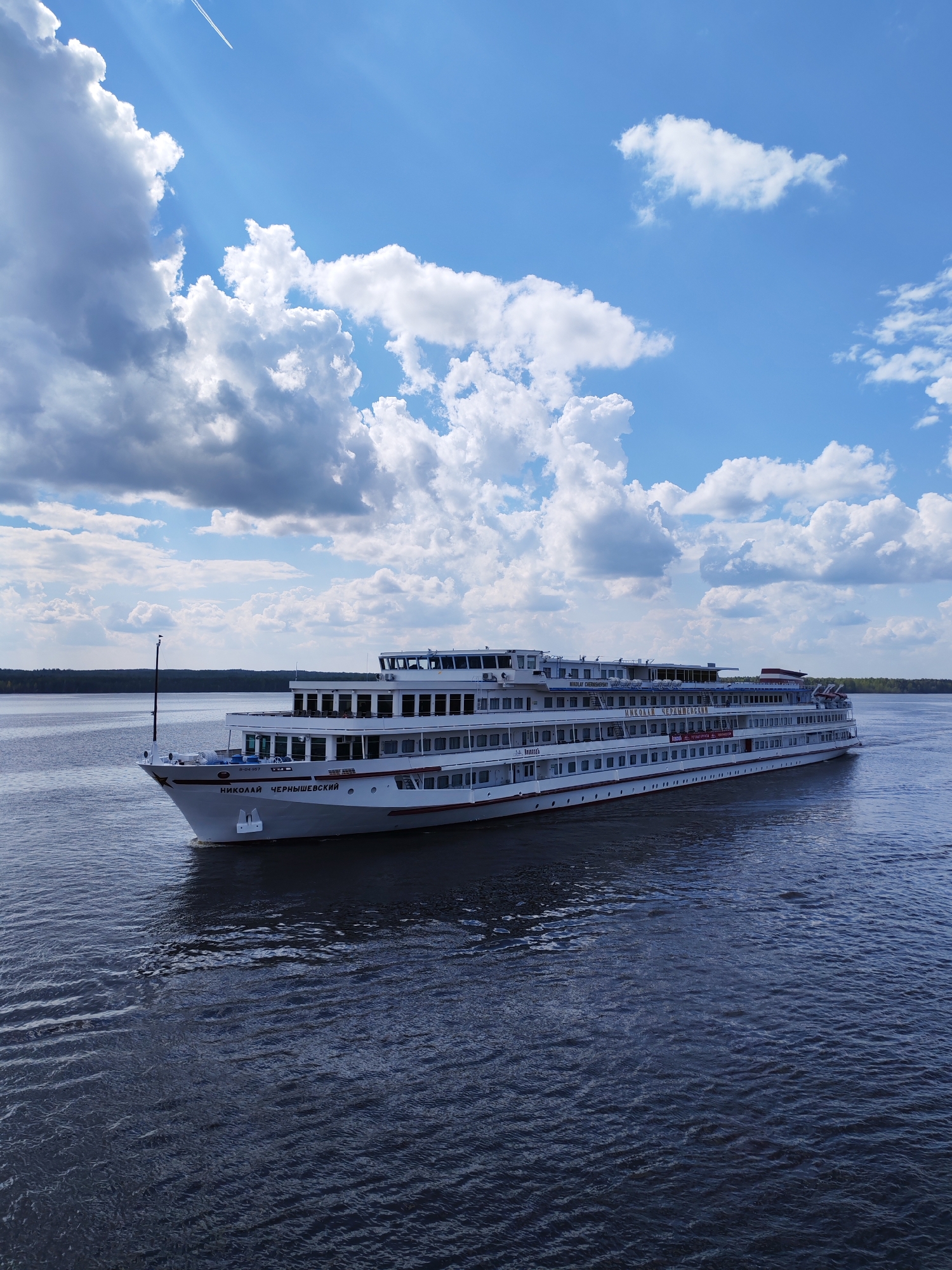 The image depicts a white, multi-deck cruise ship, named "Nikolai Chernyshevsky" in Cyrillic script on its hull, sailing on a dark blue body of water. The ship is oriented towards the left of the frame. In the background, a thin strip of land with dark green trees is visible. The sky is bright blue with large, fluffy white clouds and contrails of an airplane. The water shows subtle ripples and reflections of the sky. The location is most likely Pruzhinino, Russia, given the Cyrillic writing on the ship.