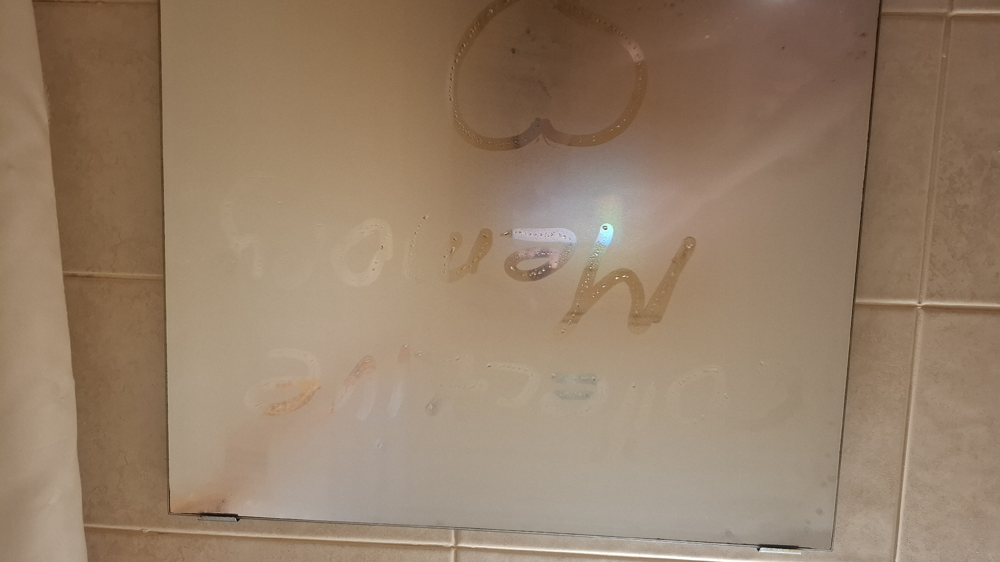 The image displays a steamy bathroom mirror, likely fogged from a hot shower or bath. At the top center of the mirror, a heart shape has been drawn in the condensation. Below the heart, two lines of text are visible, also wiped into the steam. The top line of text appears to read "Menoy" (possibly a name), and the second line below it reads "I love". This suggests a personal message of affection.
The mirror is mounted on a light brown or beige tiled wall, visible on the right and at the bottom. The tiles have visible grout lines. On the far left of the image, a light-colored, possibly white, shower curtain is partially visible, indicating a shower or bathtub area nearby. Small metal clips or brackets secure the bottom corners of the mirror. Reflections of light sources, possibly from overhead lighting, are faintly visible on the mirror's surface, creating some colorful refractions. The scene captures an intimate moment, with the drawn heart and written message conveying sentiments of love or endearment. No specific visual cues in the image directly indicate the location as Saint Petersburg, Russia.