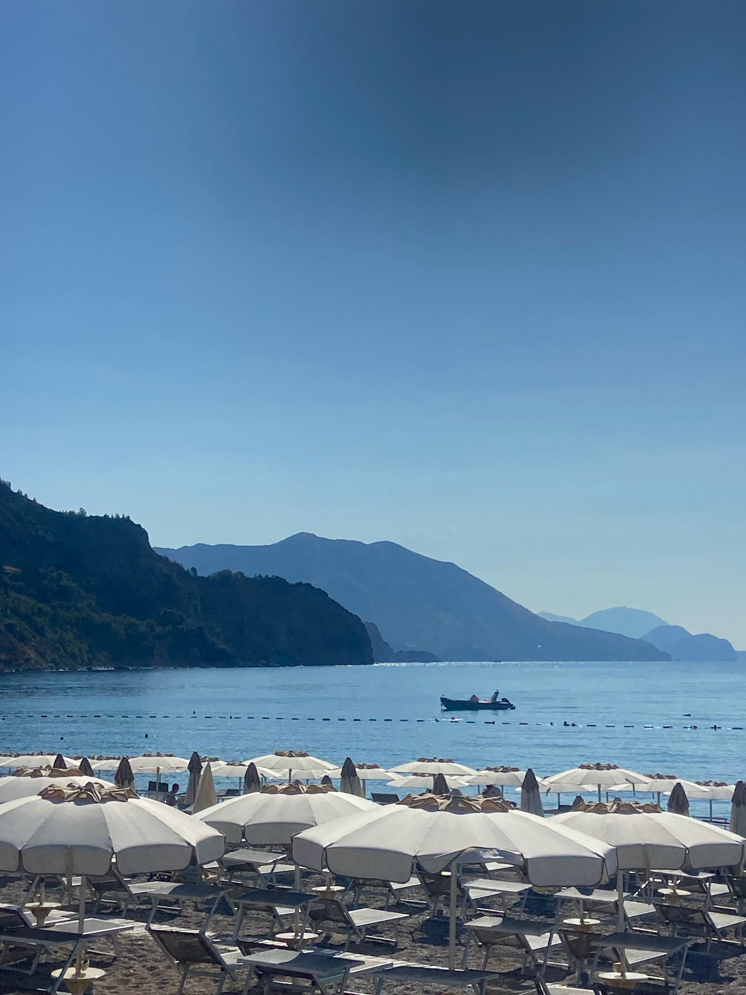 The image depicts a beach scene in Prijevor, Montenegro, characterized by a clear, pale blue sky dominating the upper portion of the frame. Below the sky, a calm, deep blue sea stretches towards a mountainous coastline, rendered in muted blues and greens due to atmospheric perspective. The foreground is a beach covered in sand, populated with rows of white beach umbrellas and loungers, most of which are unoccupied. A small boat with at least two people aboard is visible on the water, further offshore. The overall impression is one of a serene, sun-drenched day at the seaside.