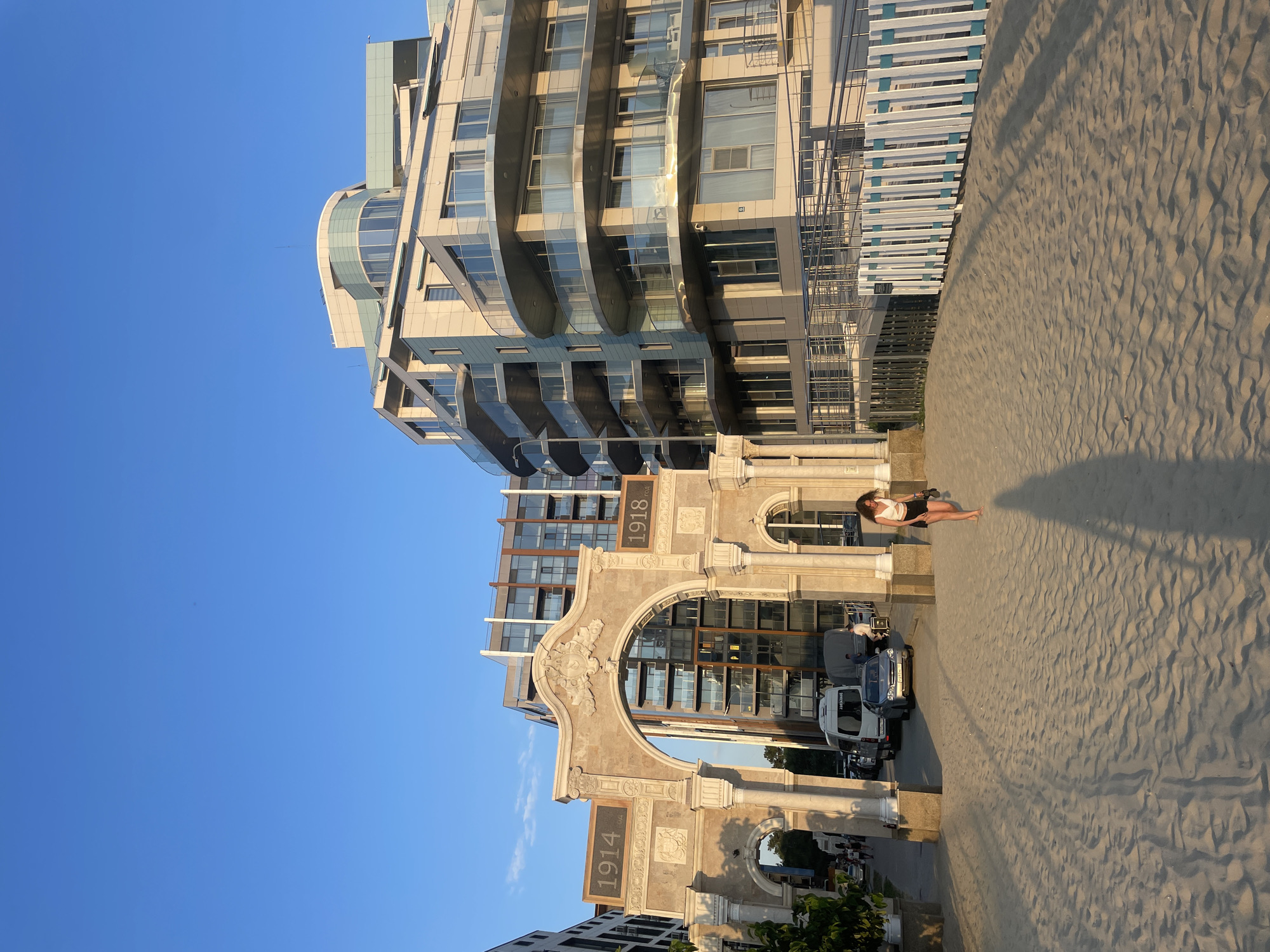 The image shows a sandy area in front of a modern building with a glass facade. A woman in a white top and black shorts is lying on the sand, facing away from the camera. Behind her is a historical arched structure with the numbers "1918" and "1914" inscribed on it. Several vehicles are parked in front of the archway. The scene appears to be set in the late afternoon or early evening, as indicated by the long shadows cast across the sand. The sky is clear and blue, suggesting pleasant weather. The architectural styles of the modern building and the historic archway suggest a location with a blend of old and new. The presence of the beach and the water in the background (partially visible) suggest a coastal setting. The overall mood of the image is relaxed and serene.