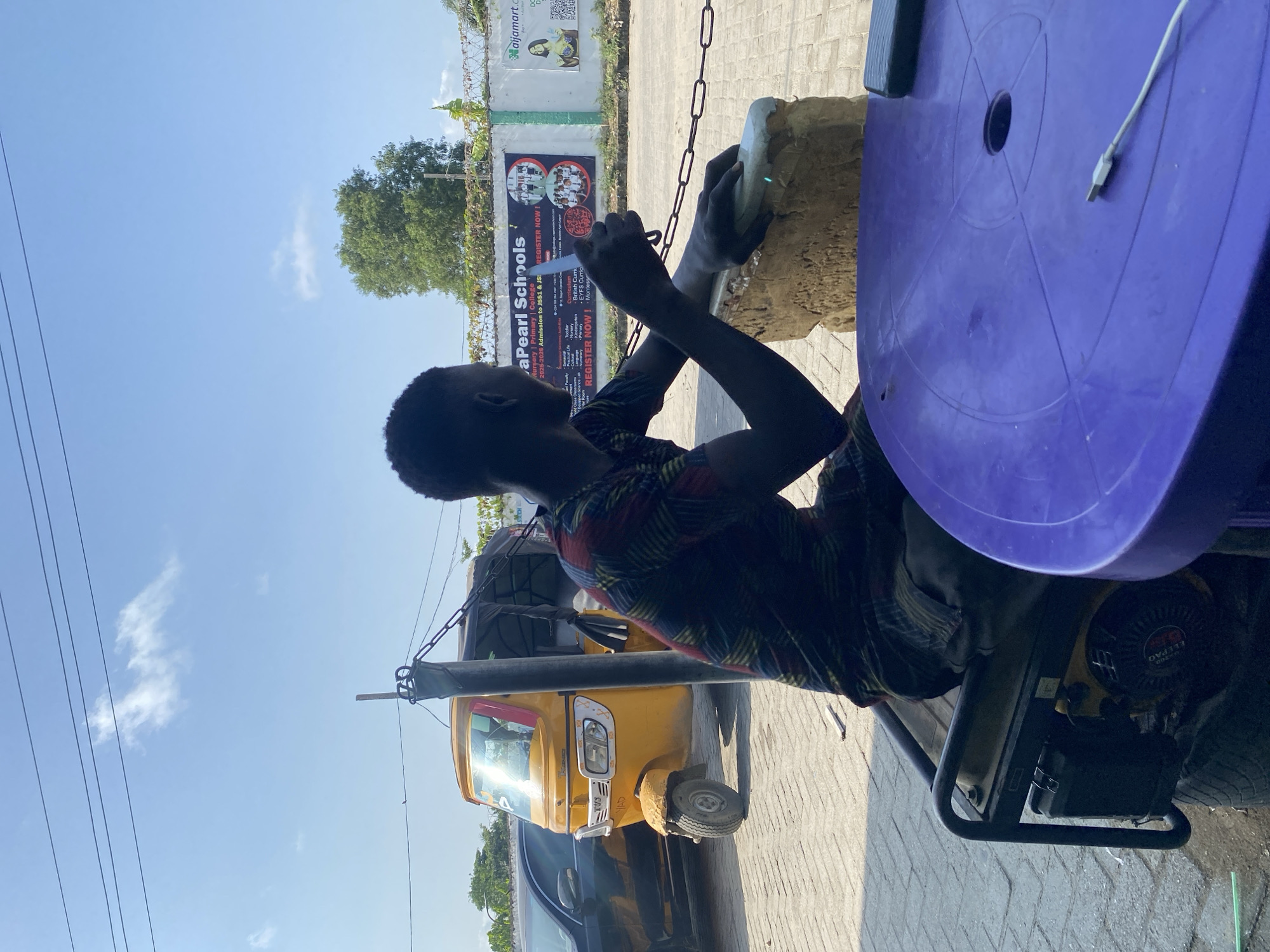 The image captures an outdoor scene in Ado, Nigeria, on a clear, sunny day. The primary subject is a person, likely a young male, seen from behind and to the side. He has dark skin and is wearing a patterned short-sleeved shirt. He is intently focused on an object held in his hands, possibly preparing or working on it. His posture suggests he is either sitting or leaning over a piece of equipment, likely a small generator visible to his right, which has "ELEPAQ" and "TVS" text on it.
To his right, partially in view, is a purple plastic surface, which appears to be a table or the top of a plastic chair. A white USB cable is lying on this surface. In the middle ground, behind the person, a bright yellow auto-rickshaw, commonly known as a Keke napep in Nigeria, is parked next to a black vehicle on a paved surface.
The background features a light blue sky with a few wispy clouds, power lines stretching across the upper left, and green trees. A white wall runs across the mid-background, adorned with two large advertisements. One prominent sign advertises "aPearl Schools," detailing "2025-2026 Admission to JSS1 & JS3," mentioning "Nursery | Primary | College," "British Curriculum," "EYFS Curriculum," and "Montessori," with a "REGISTER NOW!" call to action. Another smaller sign on the wall shows the logo "naijamart.com." The overall impression is one of everyday life in a bustling outdoor environment.