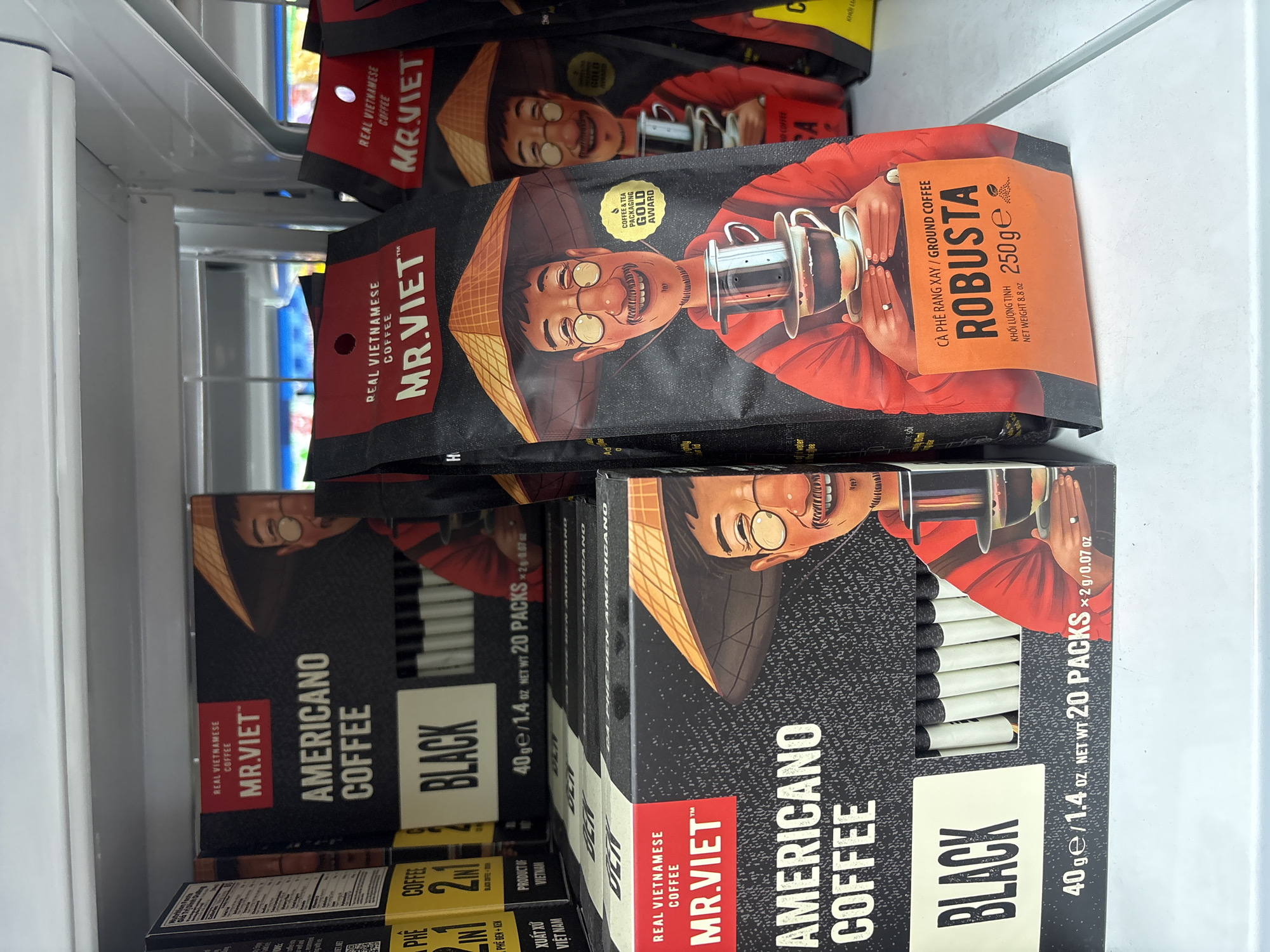 The image shows a retail display of Mr. Viet coffee products, likely in a supermarket or convenience store in Da Nang, Vietnam. The foreground features several packages of Mr. Viet Americano Coffee in black boxes and bags. One prominent black box, positioned on the lower left, displays "AMERICANO COFFEE" and "BLACK" in large white lettering. Next to it, a bag of Mr. Viet Robusta ground coffee is visible, with text in Vietnamese and English, indicating a 250g net weight. The packaging features stylized illustrations of men wearing conical hats and holding coffee-making equipment. In the background, more boxes and bags of Mr. Viet coffee are stacked on shelves. The overall environment suggests a commercial setting with brightly lit shelves and other products visible in the distance.