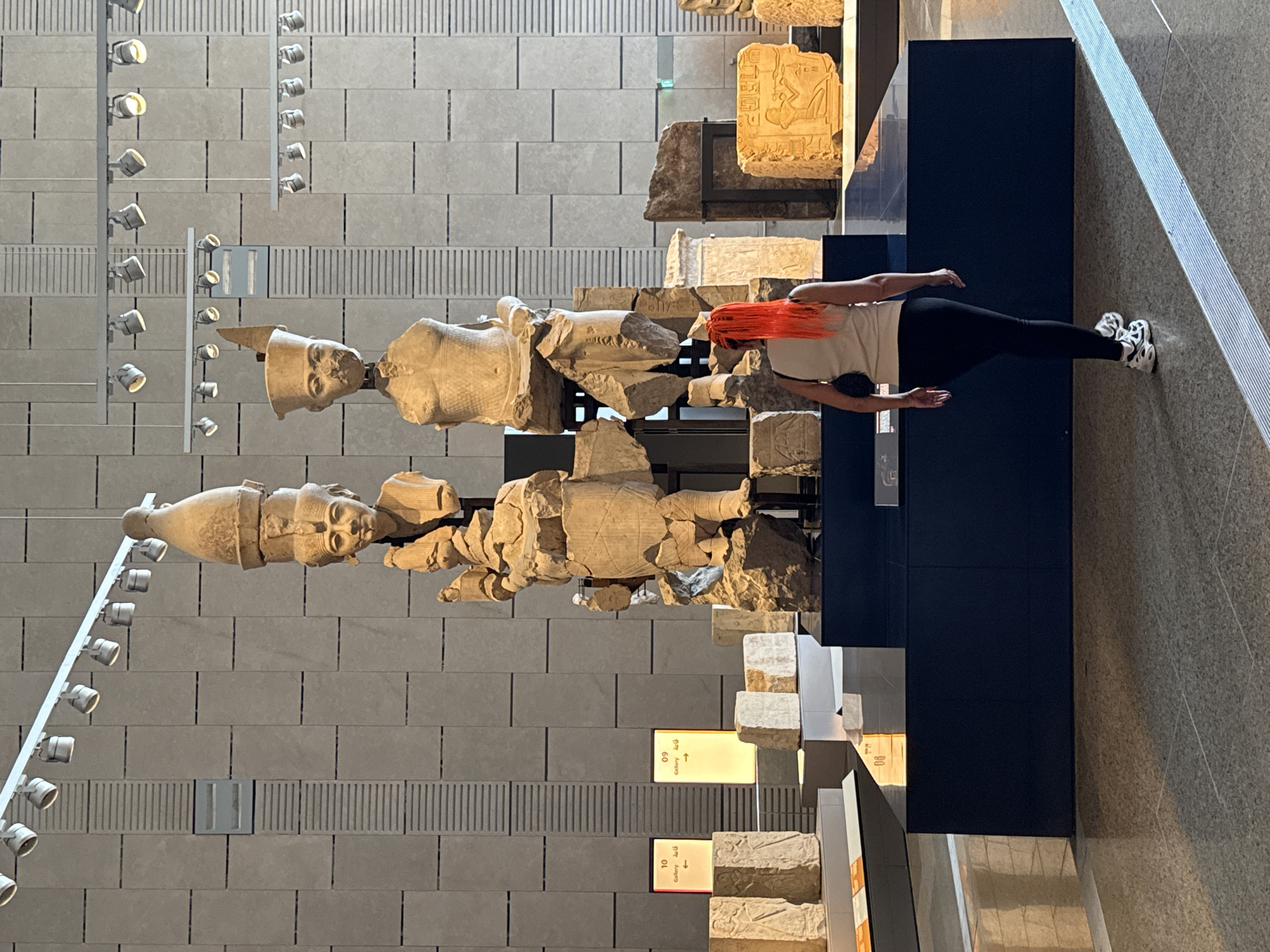 A woman with bright orange hair stands in a museum, facing away from the camera. She is wearing a cream-colored tank top, black leggings, and athletic shoes. The museum displays large, ancient Egyptian stone sculptures, including several headless figures and carved reliefs. The walls are tiled with a neutral gray pattern, and track lighting illuminates the exhibits. In the background, signs indicate gallery numbers. The floor is a polished gray surface reflecting the lights and sculptures. The image appears to be taken inside a museum, possibly a historical or archaeological exhibit, but its specific location within Kafrat Nassar, Egypt cannot be determined from the visual information provided.