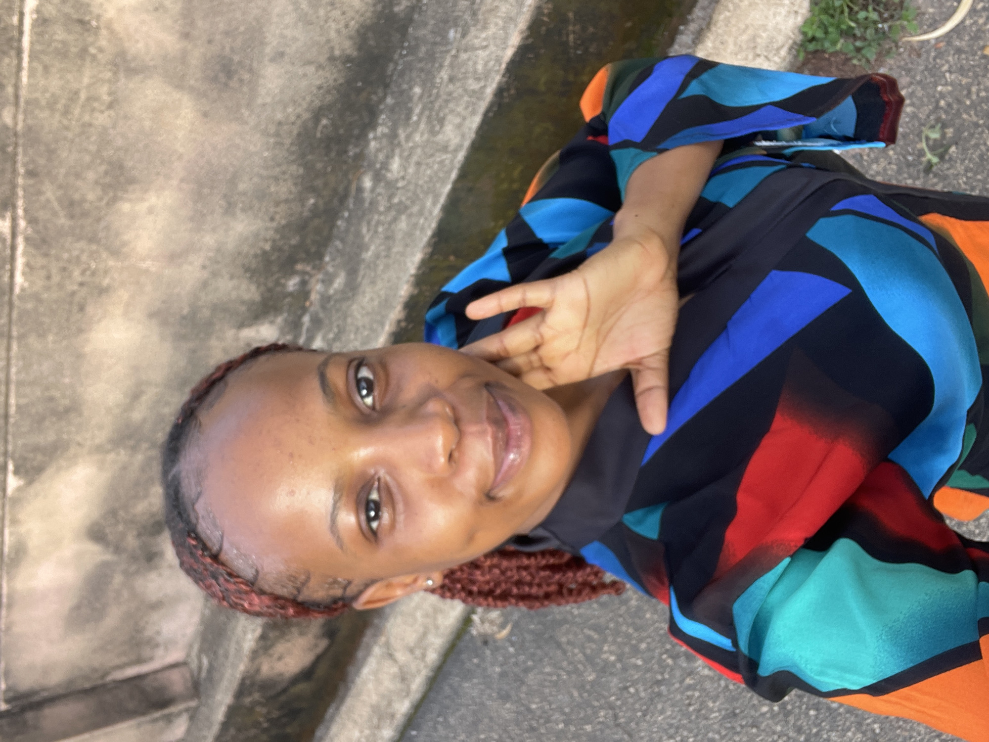 A young Black woman with braided hair and a colorful abstract print dress poses for a selfie outdoors. She is smiling and her right hand is raised to her face. The background consists of textured concrete steps and a paved surface. The lighting suggests it is daytime, possibly late afternoon. The location appears to be an outdoor public space, consistent with a location in the city of Jos, Nigeria.
