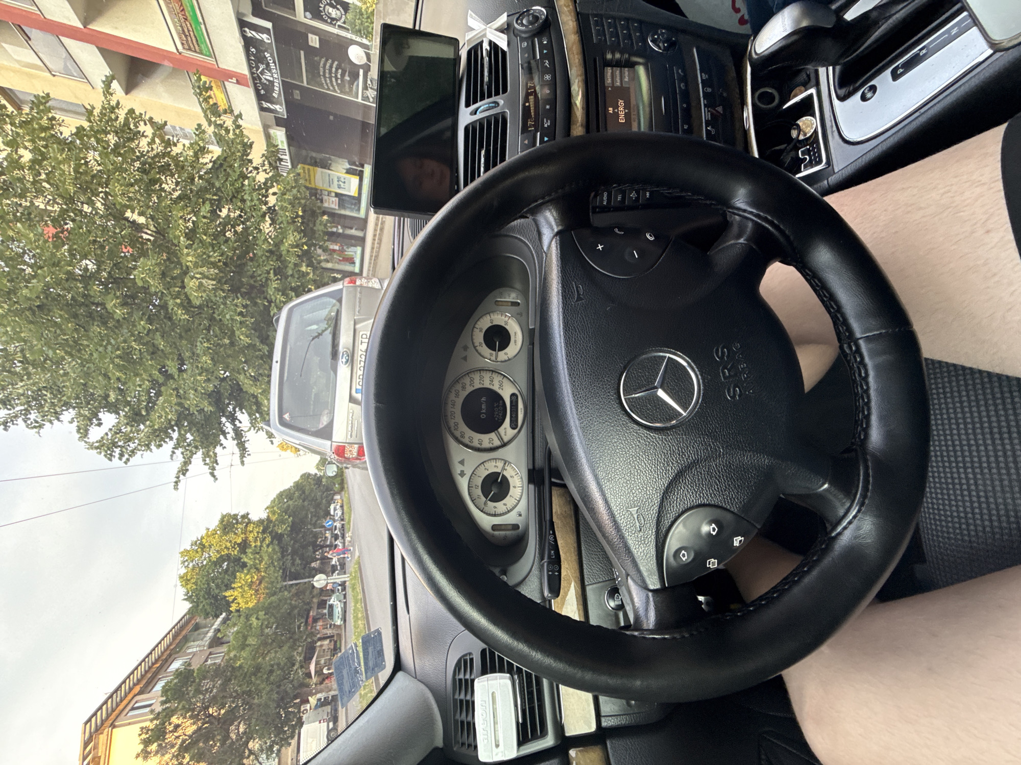 The image is a first-person view from the driver's seat of a Mercedes-Benz car, likely in Stara Zagora, Bulgaria. The car is in motion on a city street, with buildings and trees lining the road. The dashboard shows the car's instruments, including a speedometer indicating 0 km/h and an outside temperature of 29°C. A GPS device is mounted on the dashboard, displaying a reflection of a person. The steering wheel has controls for audio and phone functions. The center console shows the gear shifter and other controls. Outside the car, other vehicles, pedestrians, and shops are visible, including a barbershop with a sign reading "SOLIKOV'S BARBERSHOP". The sky is overcast with a few trees visible, suggesting it is daytime. The overall impression is a snapshot of daily life in a Bulgarian city.