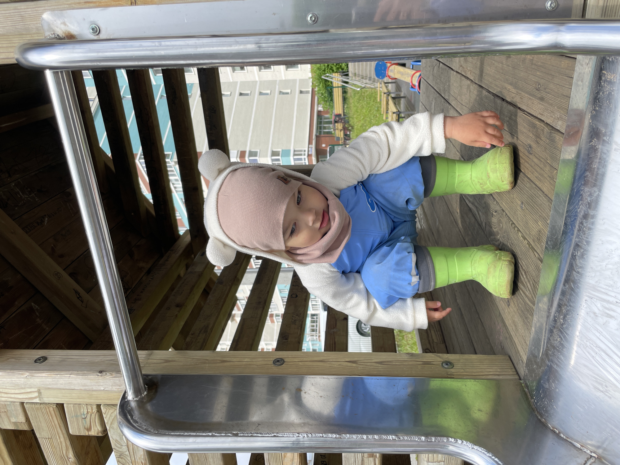 The image captures a young child positioned at the entrance of a metal slide on a wooden playground structure in Izhevsk, Russia. The scene appears to be set outdoors on a bright, sunny day.
The subject is a toddler, dressed in a white fleece-like sweater, light blue overalls or pants, and distinctive lime green rubber boots. A soft pink knitted hat with white, fuzzy ear-like decorations covers their head. The child's face is visible, showing a neutral to slightly curious expression as they look downwards and to the left. Their hands are resting on the wooden platform and the edge of the slide.
The playground structure consists of robust wooden planks and railings, forming a platform for the slide. The slide itself is made of shiny metal. In the background, a multi-story apartment building with numerous windows and a light-colored facade is prominent, indicating an urban or residential environment. Beyond the building and playground, patches of green grass and other playground equipment in the distance (featuring blue and red elements) are faintly visible.
The activity appears to be the child preparing to go down the slide or having just reached the top. The lighting suggests it is daytime with clear weather. No legible text is visible on any objects or clothing.