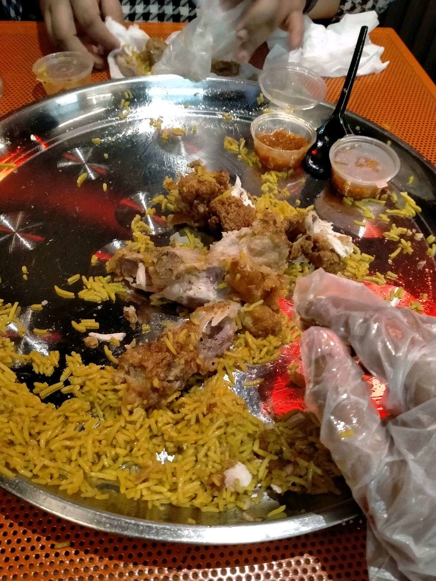 A close-up, top-down shot shows a large, round, metal platter filled with food, primarily yellow rice and pieces of fried chicken. A hand, wearing a clear plastic glove, is visible in the lower right, reaching towards the food. Two small plastic containers with dipping sauce are placed on the platter, along with a black plastic spoon. In the background, out of focus, are more hands and white paper wrappers. The platter rests on an orange, perforated placemat, which is on top of a table. The overall scene suggests a meal that has been partially consumed.