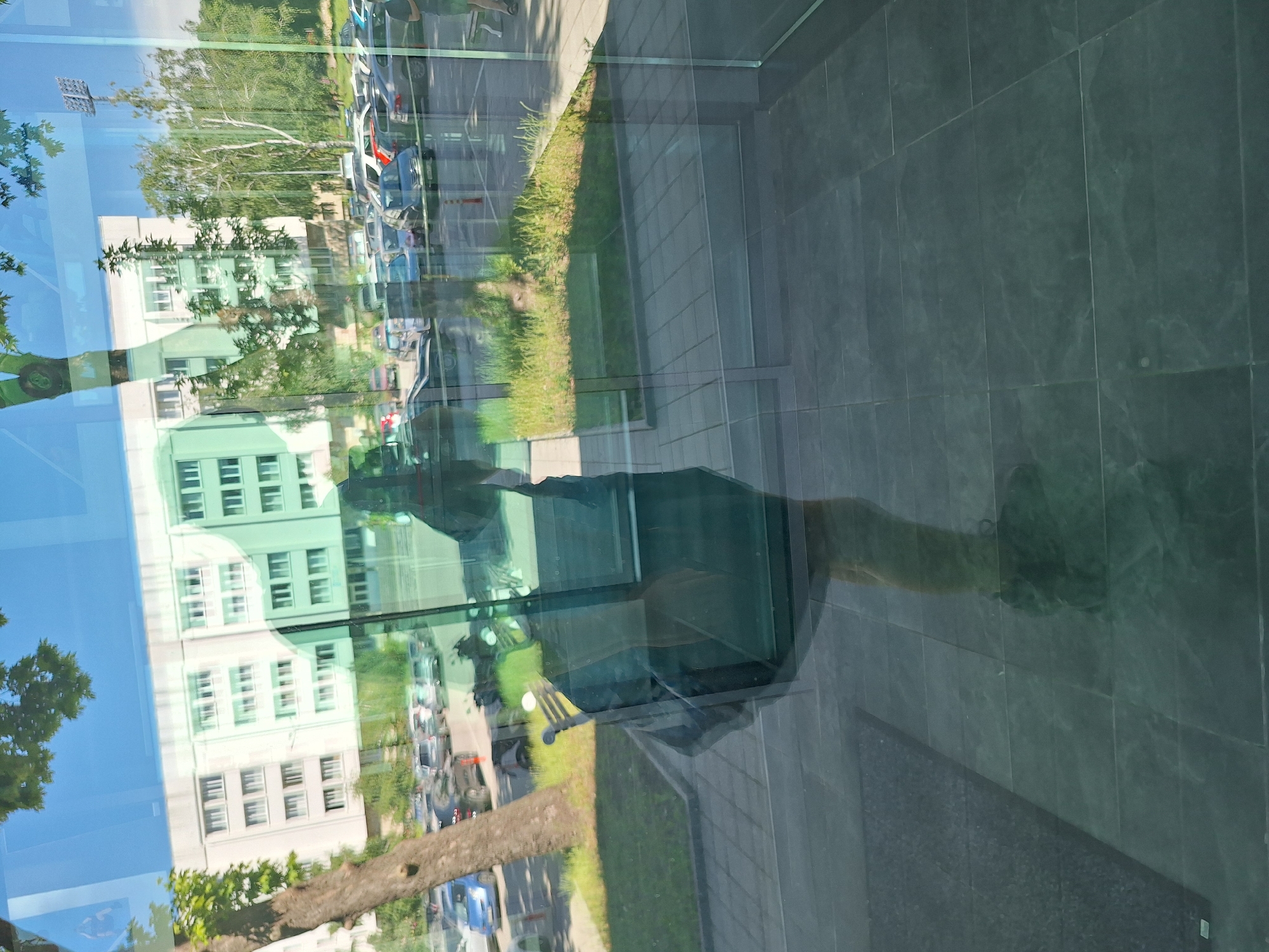 This media file captures a reflection in a large glass surface, likely a building facade. The scene depicted is an outdoor environment. In the reflection, we see a building with light-colored walls and multiple windows, suggesting it is a multi-story structure. Trees with green foliage are visible, along with a clear blue sky, indicating it is daytime and likely sunny. The reflection also shows a street with parked cars and some greenery along the side.
The reflection of a person is discernible, appearing as a silhouette or faint outline against the background. The person seems to be standing or walking, with a shadow cast across the tiled ground visible in the reflection. The overall impression is that of a bright, ordinary day. The setting appears to be urban or suburban, with the combination of a building, street, and parked vehicles. There is no visible text in the image. The quality of the reflection is high, providing a clear, albeit distorted, view of the surrounding environment.