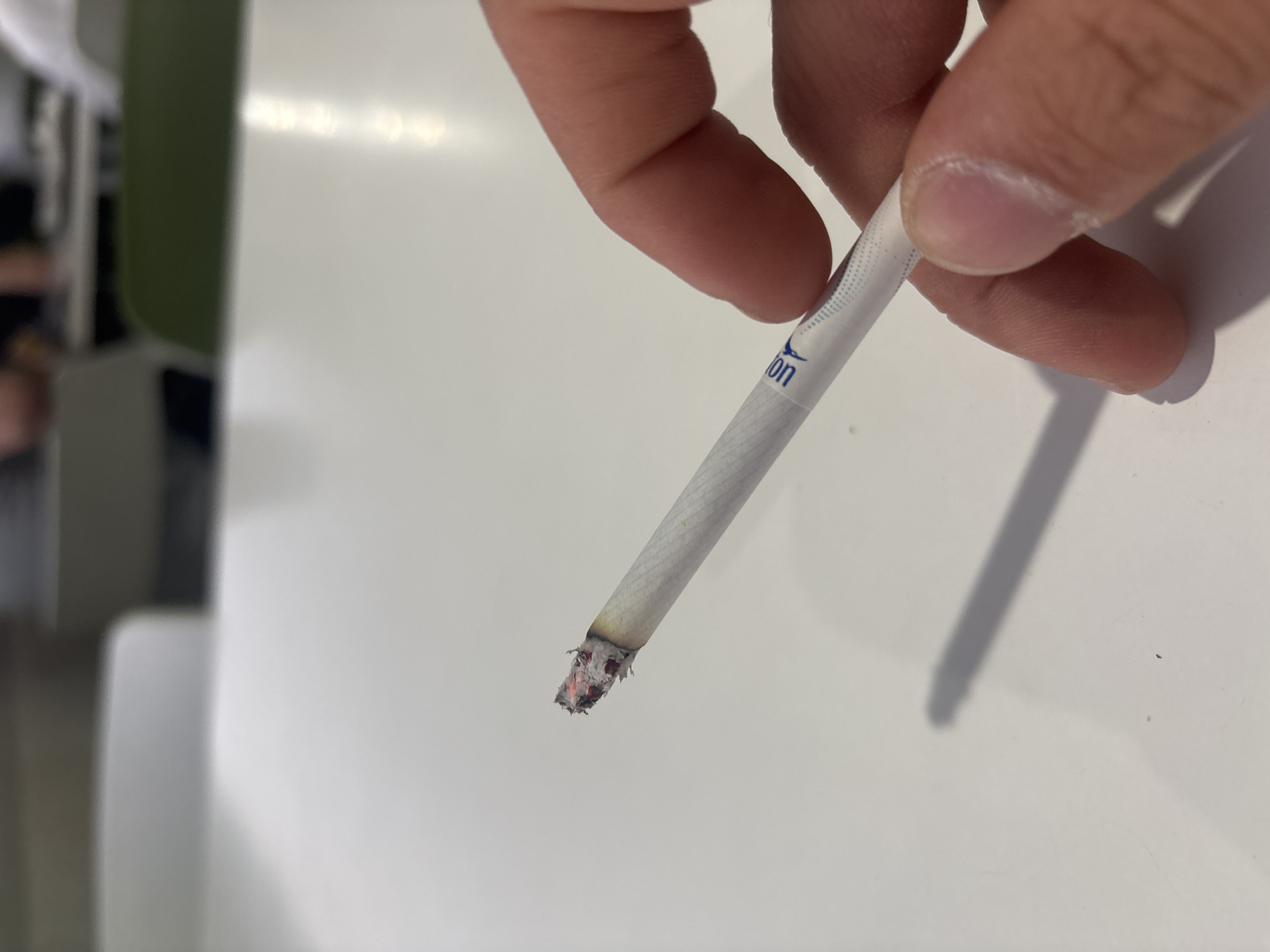 A close-up shot features a person's hand holding a lit cigarette against a bright, reflective white surface, likely a table or desk. The fingers and thumb of the hand are clearly visible, grasping the cigarette near its filter. The tip of the cigarette is actively burning, indicated by the glowing red embers and accumulated ash.
On the white filter paper of the cigarette, there is a blue graphic logo that resembles a stylized bird or an abstract winged design. Partially visible blue text "on" can also be seen next to the logo.
The background of the image is out of focus, displaying blurred shapes and muted colors, including shades of green, gray, and darker tones, suggesting an indoor setting such as a room or office. There are no explicit visual cues within the image to determine the time of day or specific weather conditions. While the provided context indicates the location is Yerevan, Armenia, this information is not visually discernable from the scene depicted. The image solely focuses on the act of holding the lit cigarette, without revealing any facial expressions or additional subjects.