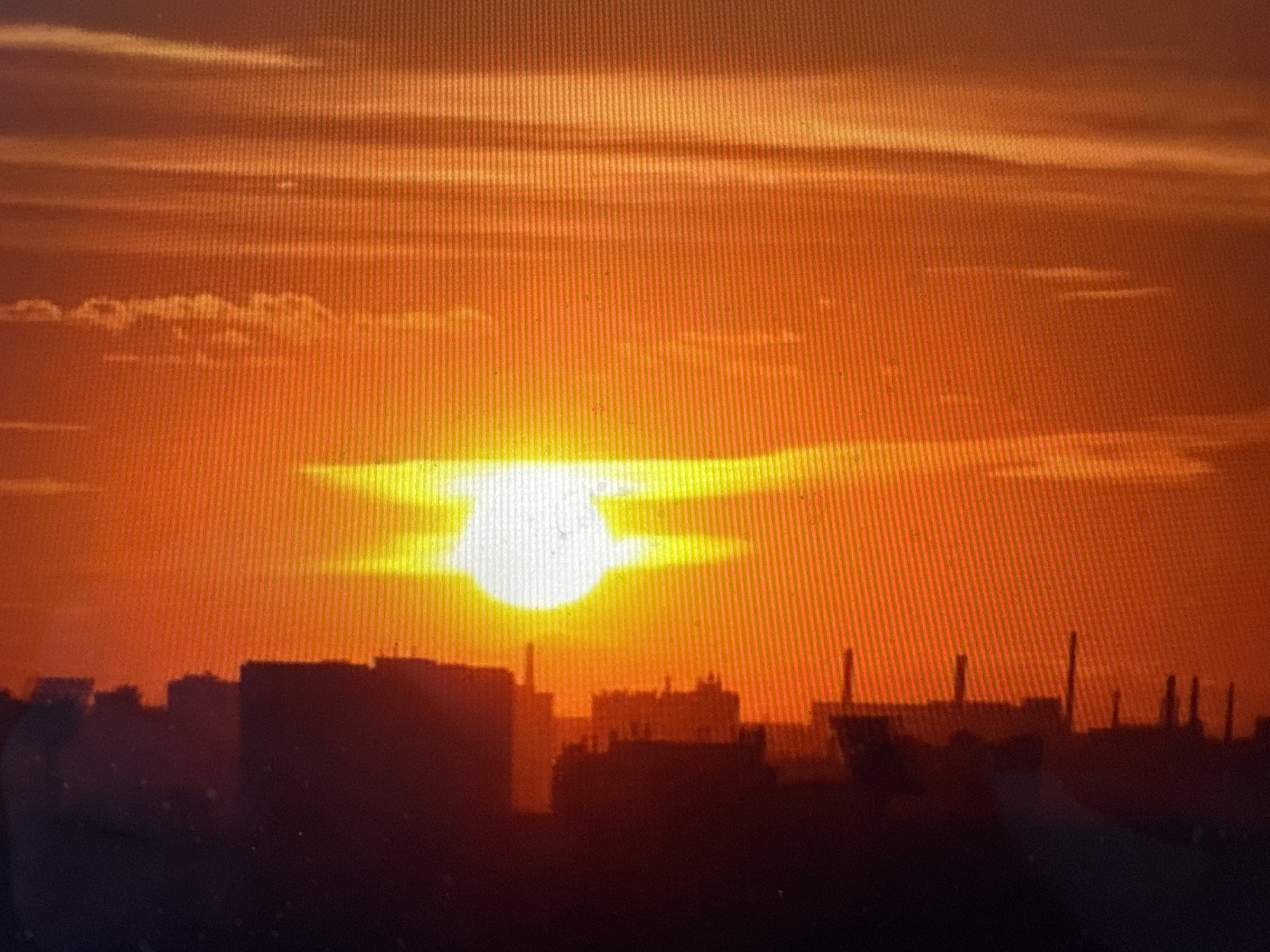 The image captures a cityscape at sunset. The sky is dominated by a vibrant orange and yellow hue, with horizontal clouds creating striations across the horizon. The sun, a bright white disk, is partially obscured by a low-lying cloud formation, giving it a flattened appearance. In the foreground, the silhouettes of various buildings and industrial structures, possibly chimneys, are visible against the dramatic sky. The overall impression is one of urban industrialization meeting a powerful natural phenomenon.