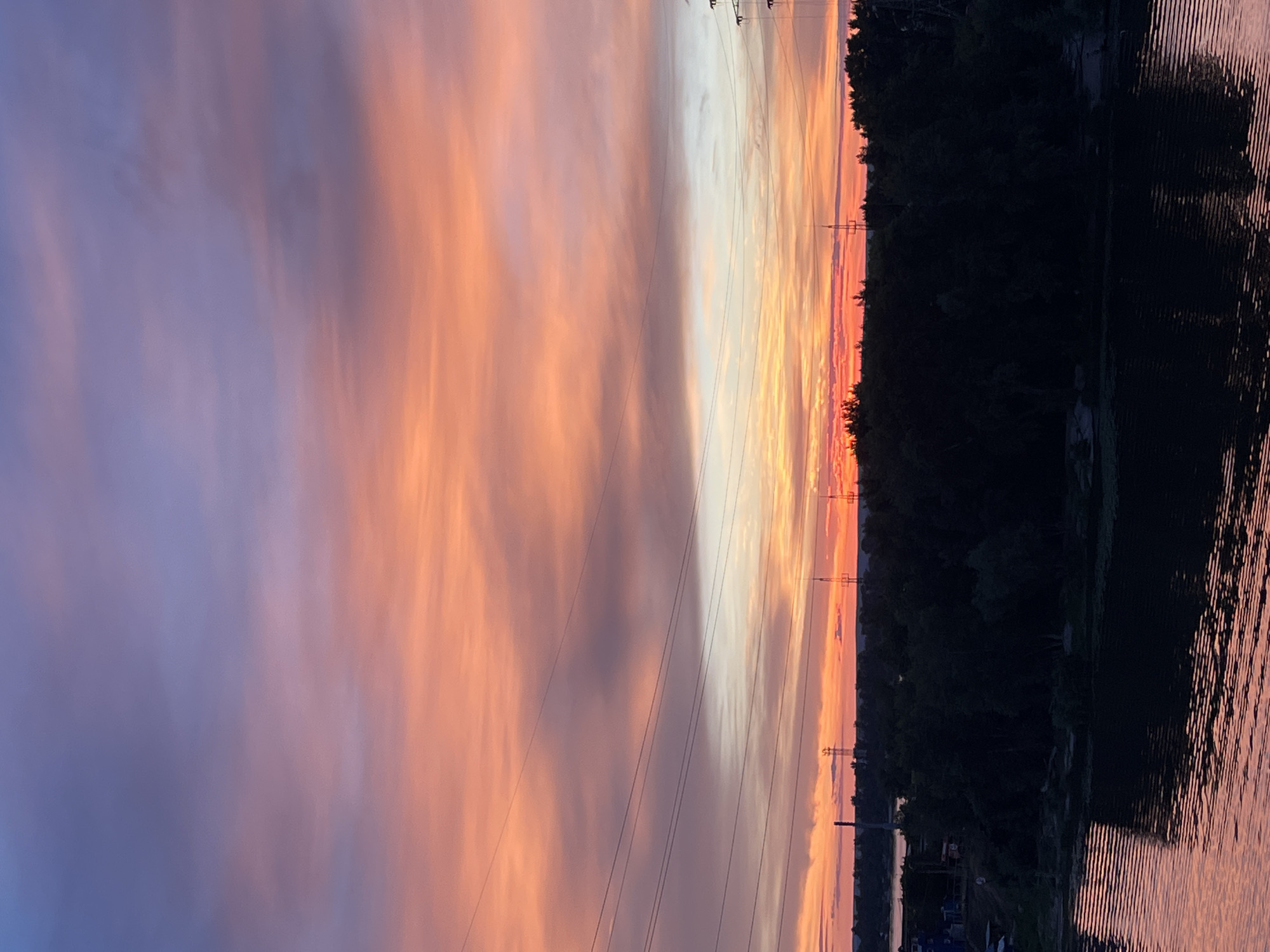 The image shows a sunset over the Volga River in Samara, Russia. The sky is filled with vibrant colors of orange, pink, and purple, with scattered clouds. In the foreground, the dark water of the river reflects the colors of the sky. A dark silhouette of a forest lines the riverbank, with several power lines crisscrossing the scene. In the distance, faint structures and lights suggest the presence of urban development along the river.
