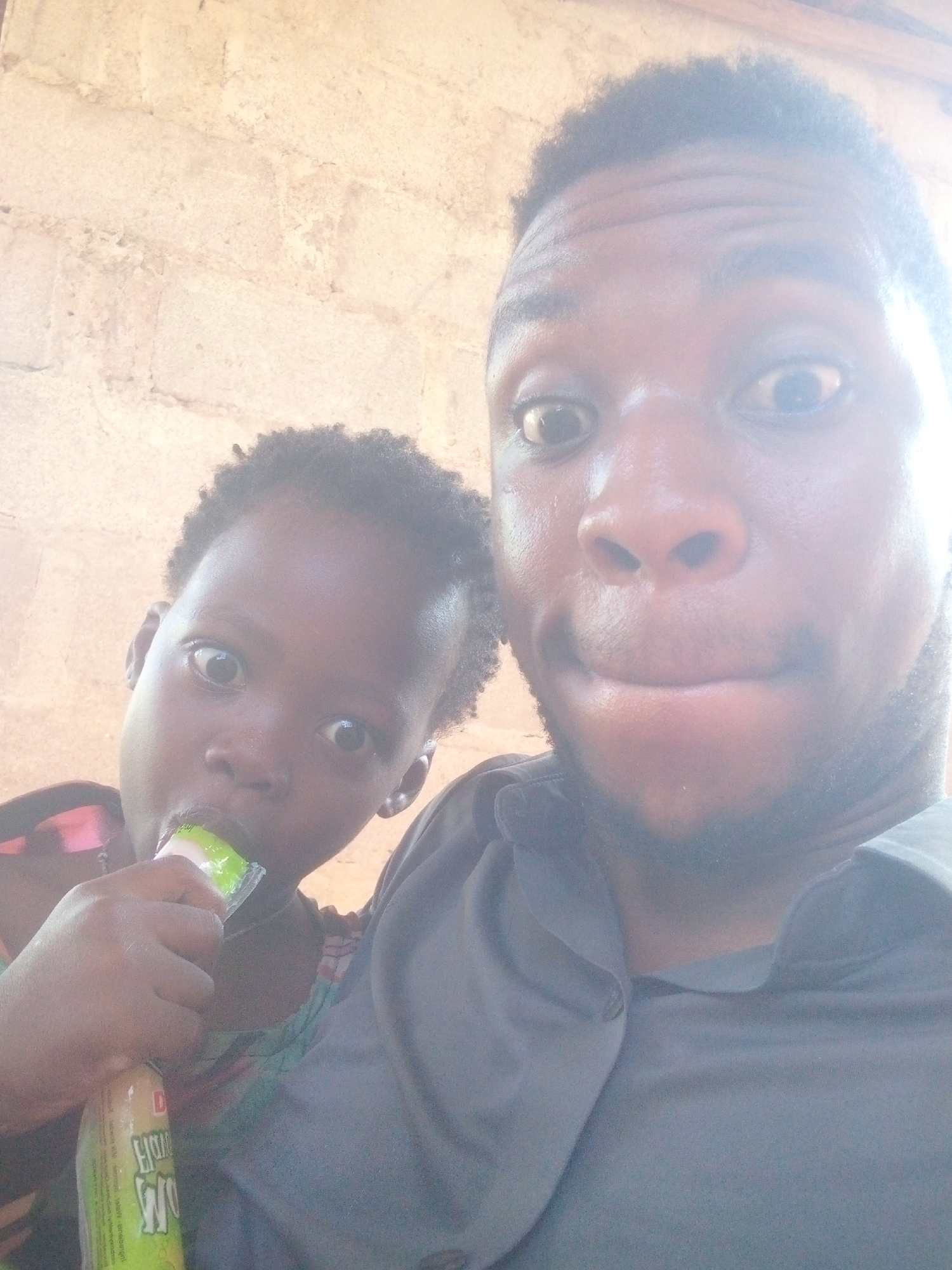 The image shows a selfie of a man and a young girl, both looking directly at the camera with slightly surprised expressions. The girl is holding a green and white beverage container to her mouth. The background appears to be an interior space with a light-colored wall. The city and specific location are unknown.