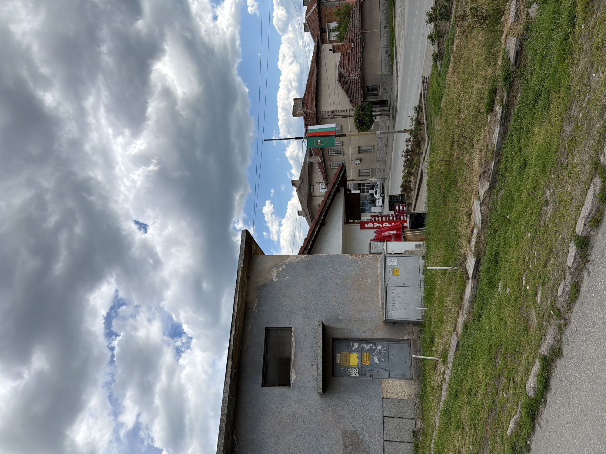 The image depicts a street scene in Panagyurishte, Bulgaria, with a focus on a small, gray, concrete building. The building features a single small window and a metal door with warning signs in Bulgarian. Adjacent to the building are utility boxes.
The Bulgarian flag is visible on a pole down the street. Other buildings line the street. A patch of green grass and stones separates the building from the road. The sky is cloudy.