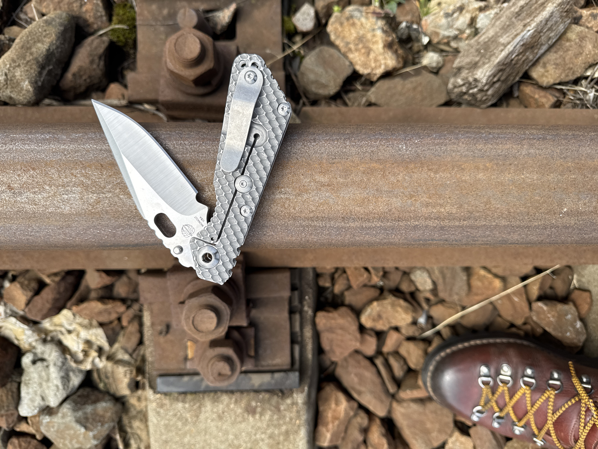 A partially open pocketknife rests on a railway track. The knife has a silver-colored blade and a textured silver handle with a clip. The railway track is rusty, with bolts and gravel surrounding it. The tip of a brown hiking boot with yellow laces is visible on the right side of the image. The location is not discernible from the image. The knife appears to be stationary on the track. There are no people visible.