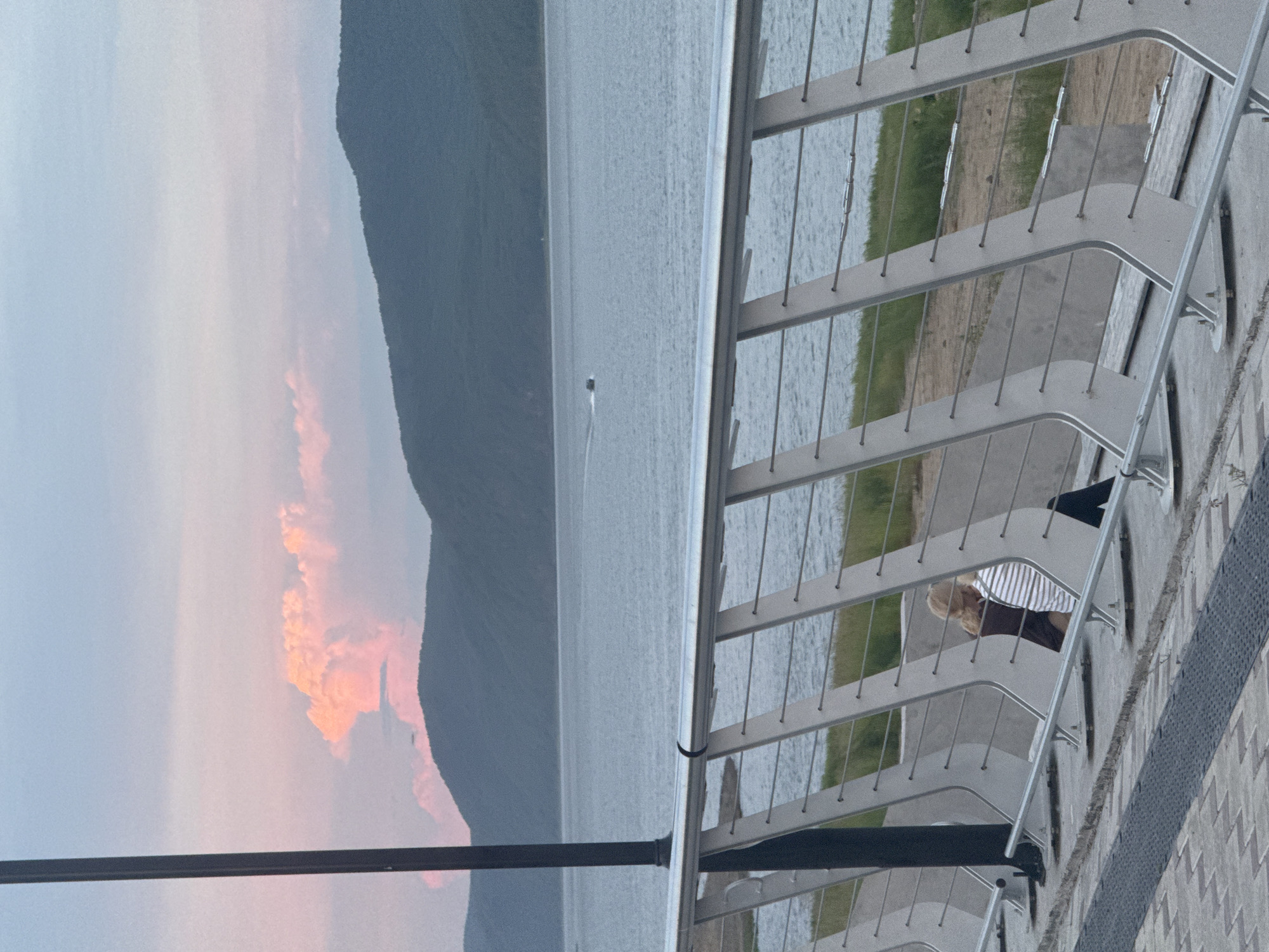 The image shows a waterfront scene, likely in Komsomolsk-on-Amur, Russia, during what appears to be twilight or early morning. The foreground features a modern metal railing with diagonal supports and horizontal wires. Two individuals are seated on a bench behind the railing. One person, with blonde hair, is wearing a dark top and striped pants, leaning towards the other person who is wearing a dark garment. To the left, a dark lamppost is visible. The middle ground consists of a calm body of water, possibly the Amur River, with a small boat or jet ski visible in the distance. In the background, dark green, tree-covered hills form a silhouette against a pale sky tinged with pink and blue clouds. The ground in the immediate foreground is a paved walkway with a textured surface.